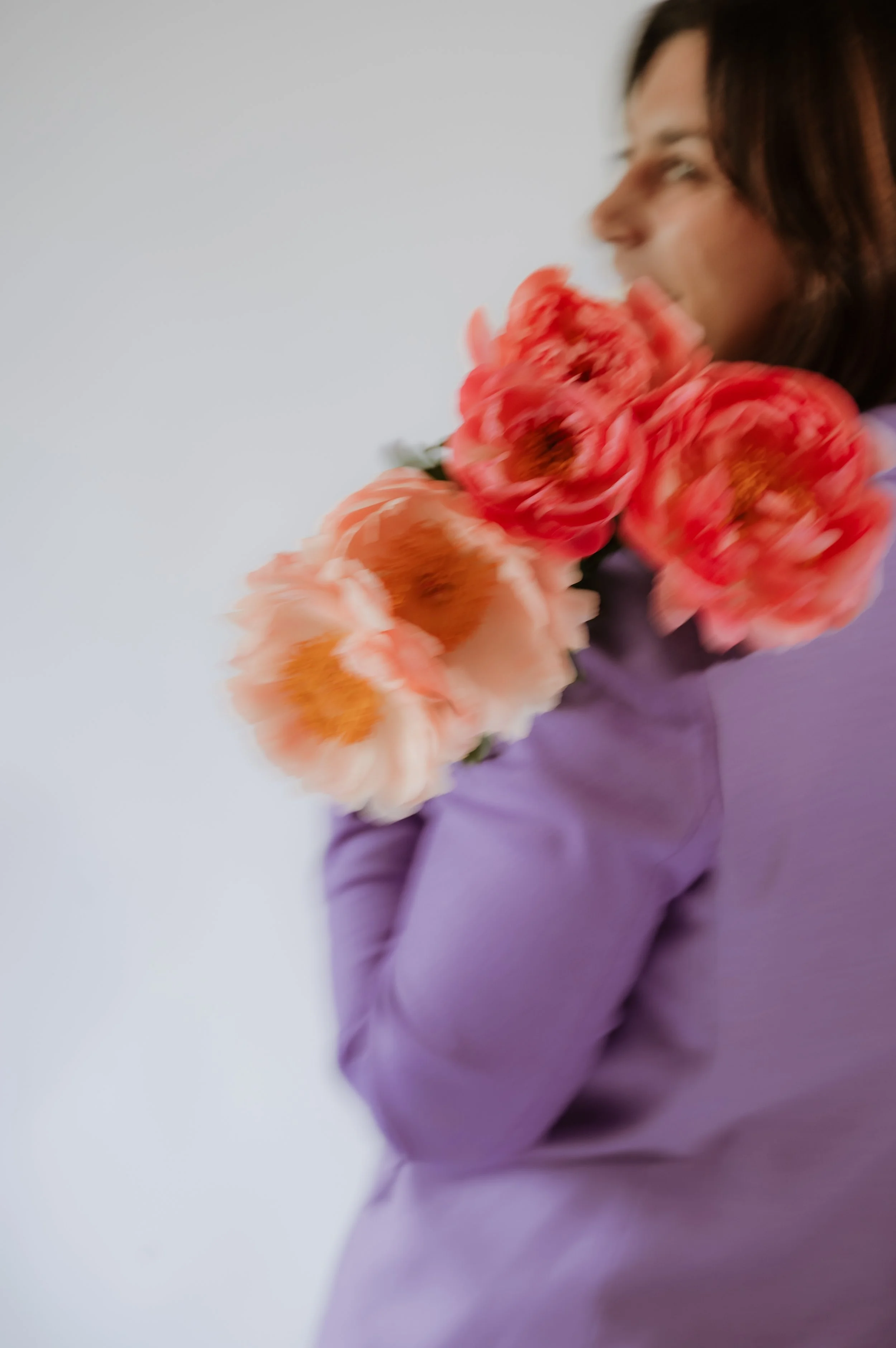 A woman with dark hair holding a bouquet of pink and orange peony flowers, with a neutral plain background.