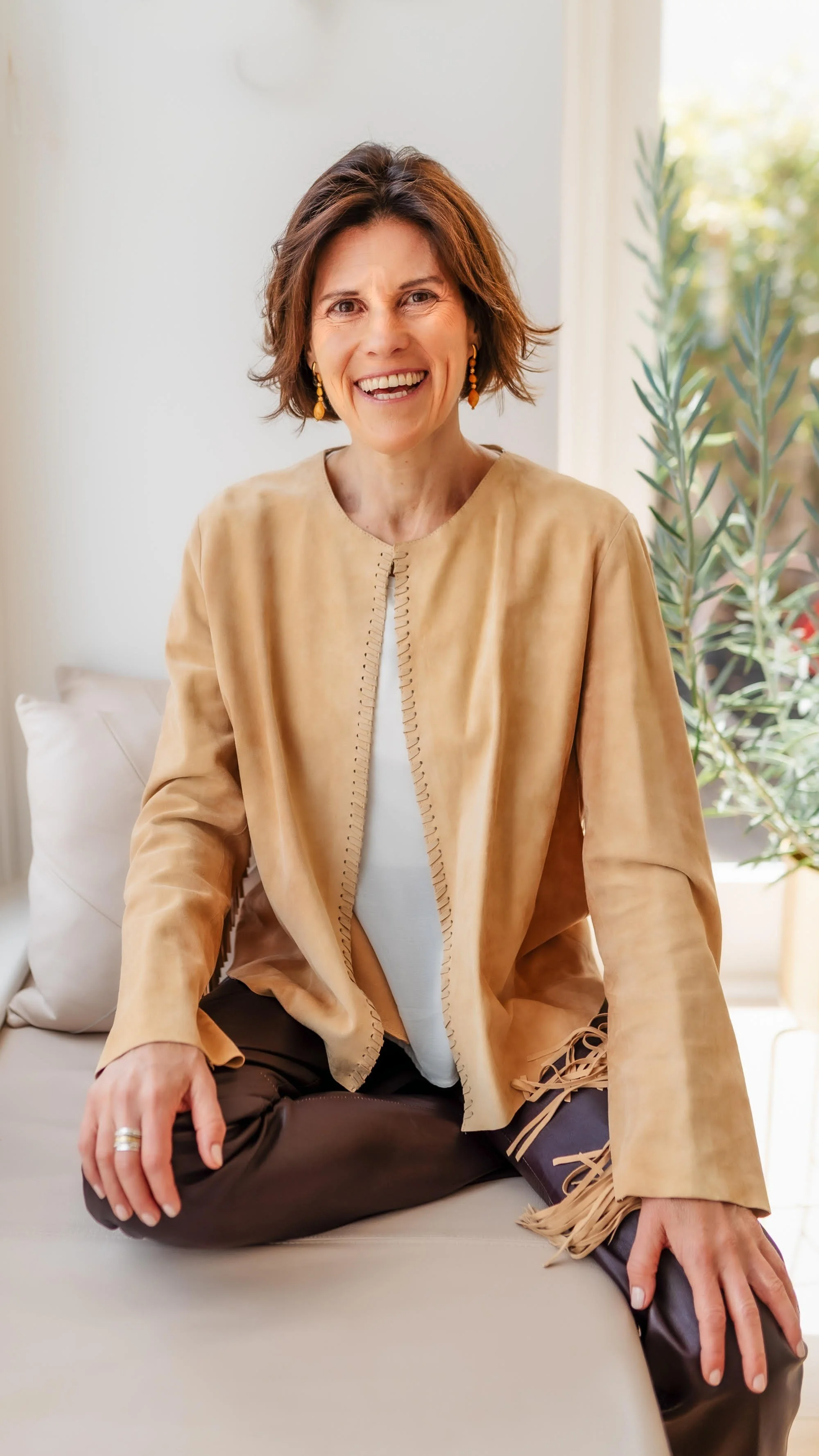 A woman sitting cross-legged on a beige couch, smiling in a light-filled room with a plant and a window in the background.