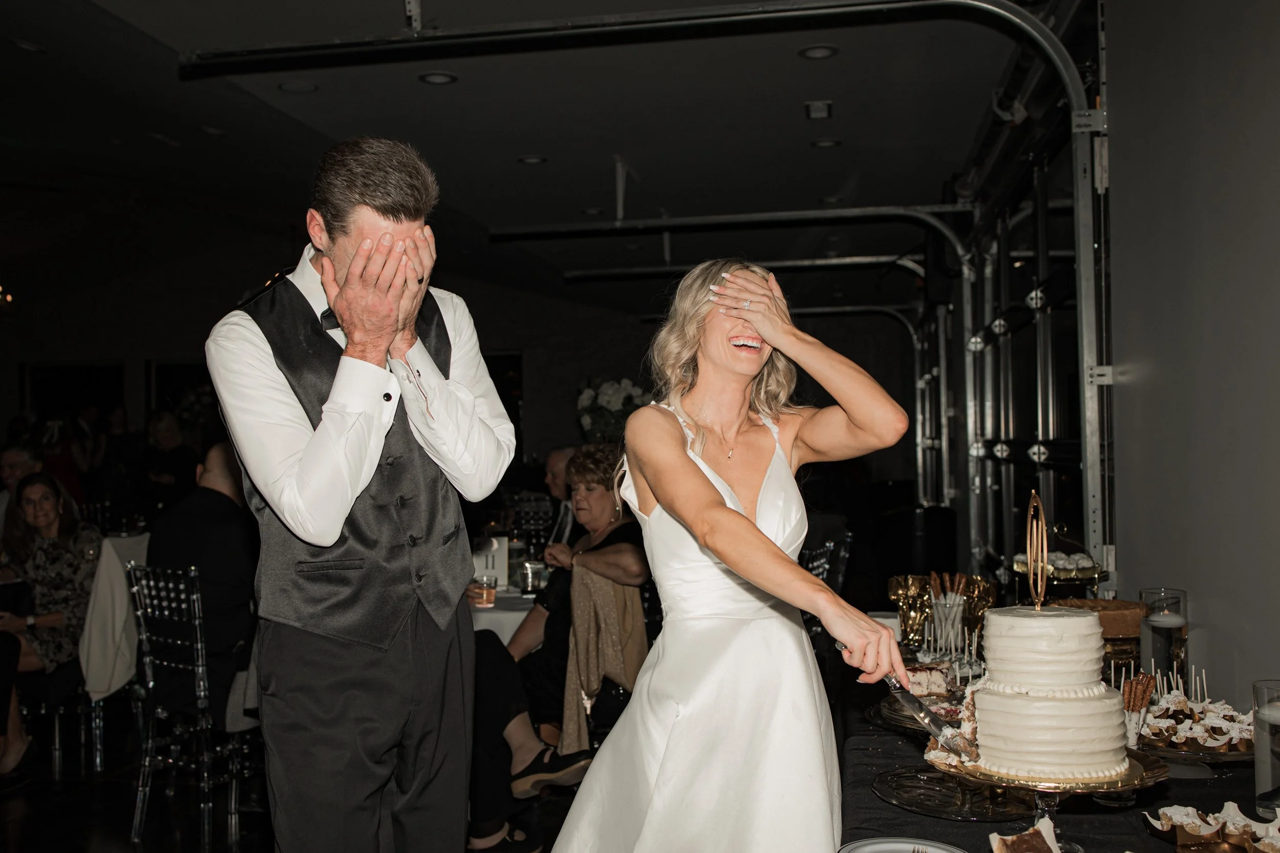 A bride and groom playfully covering their faces while cutting a wedding cake at a reception.
