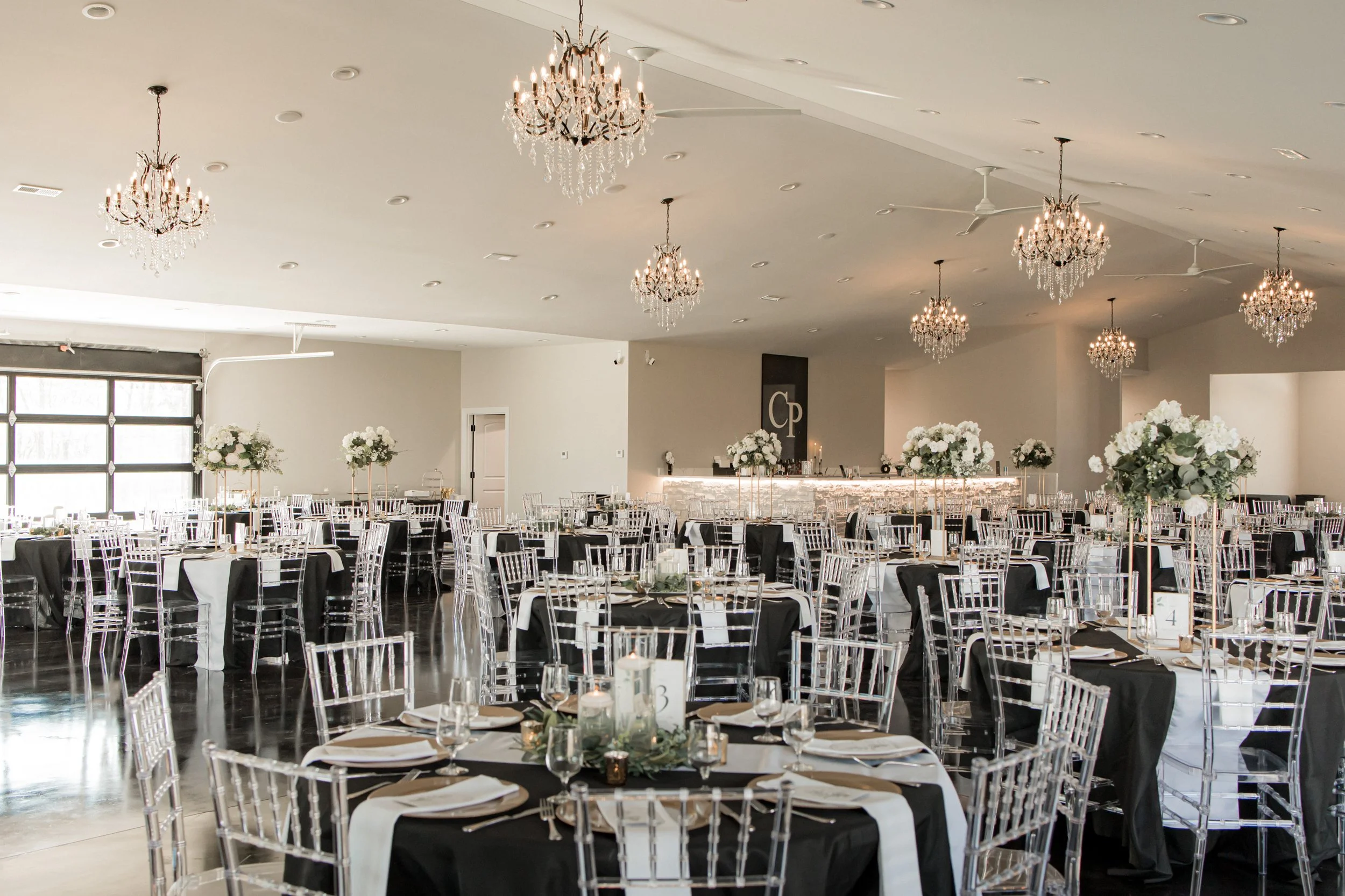 Elegantly decorated banquet hall with chandeliers, transparent chairs, round tables with black and white tablecloths, and tall floral centerpieces.