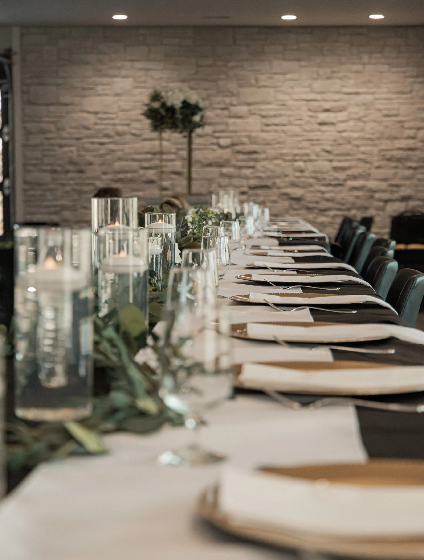 Elegant dining table setup with white tablecloths, glass candle holders, greenery, and place settings in a stone-walled room.
