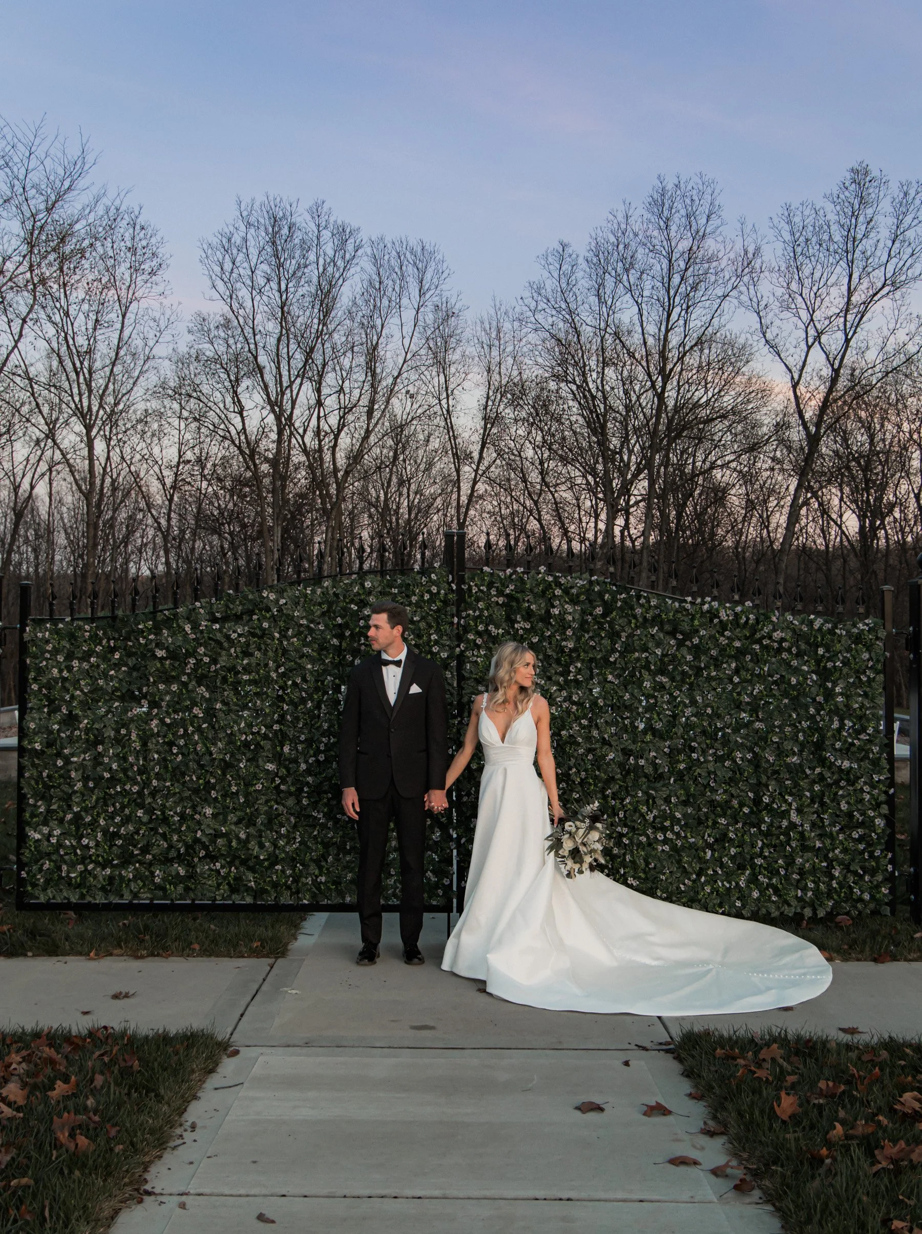 Bride and groom standing outdoors in front of a green floral backdrop, holding hands; the bride wears a white gown with a long train, and the groom wears a black tuxedo.