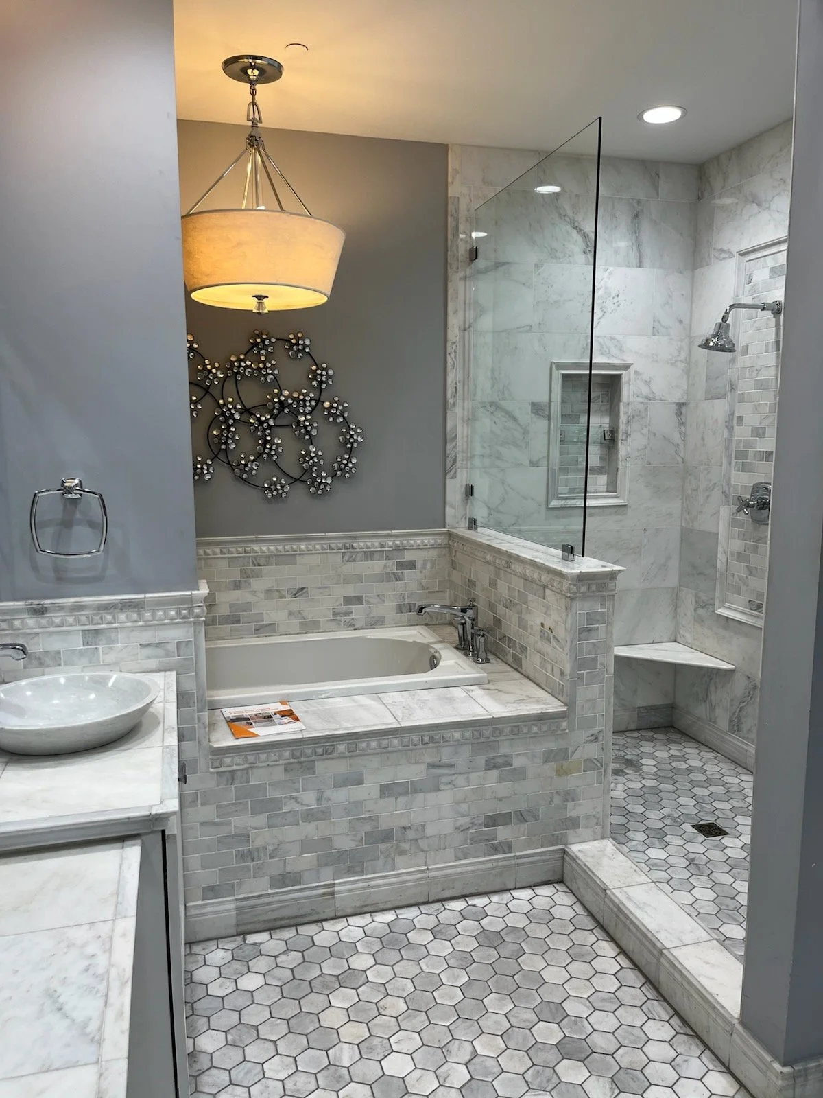 Bathroom with gray and white marble tiles, a glass-enclosed shower, a bathtub with a marble surround, a vessel sink, and a hanging light fixture.