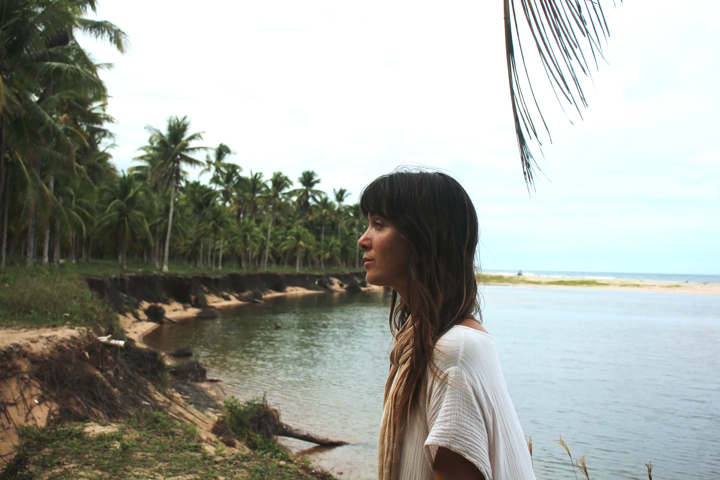 Side view of a woman with long dark hair standing near a river at the beach, with palm trees and the ocean in the background.