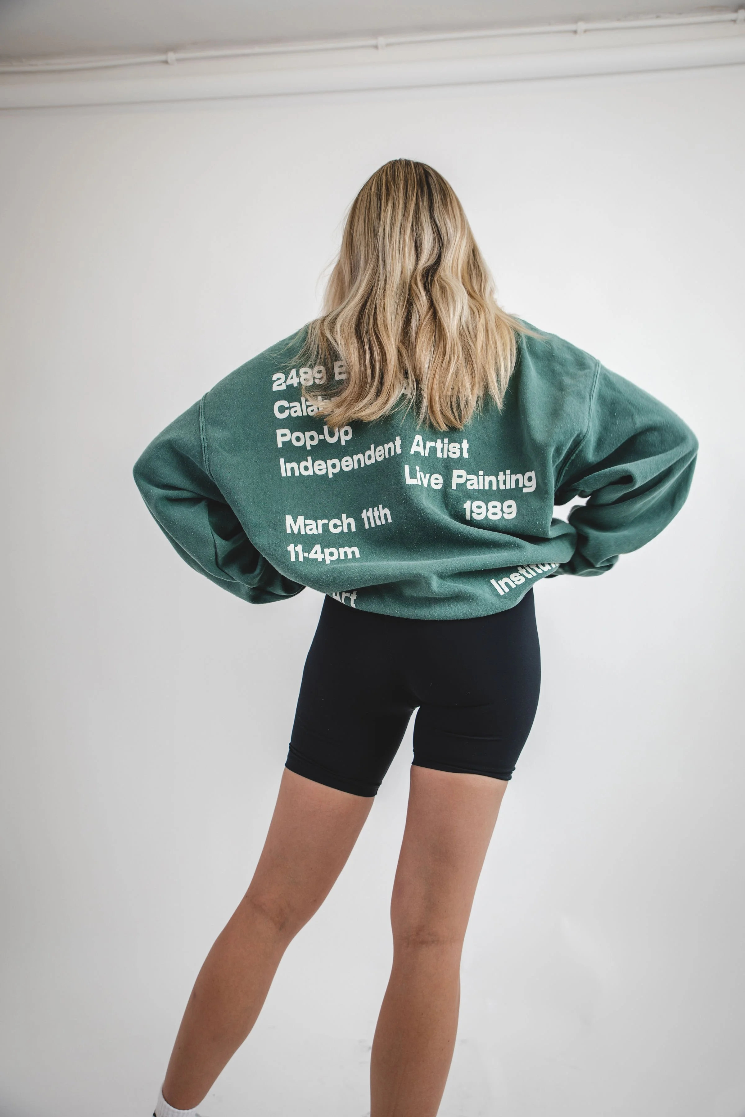 Back view of a woman with wavy blonde hair wearing a green sweatshirt and black shorts, posing with her hands on her hips in front of a plain white wall.