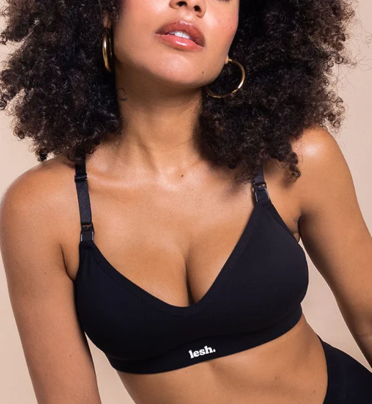 Close-up of a woman with curly hair wearing gold hoop earrings and a black bralette with the brand name 'lesh.' visible on the band.