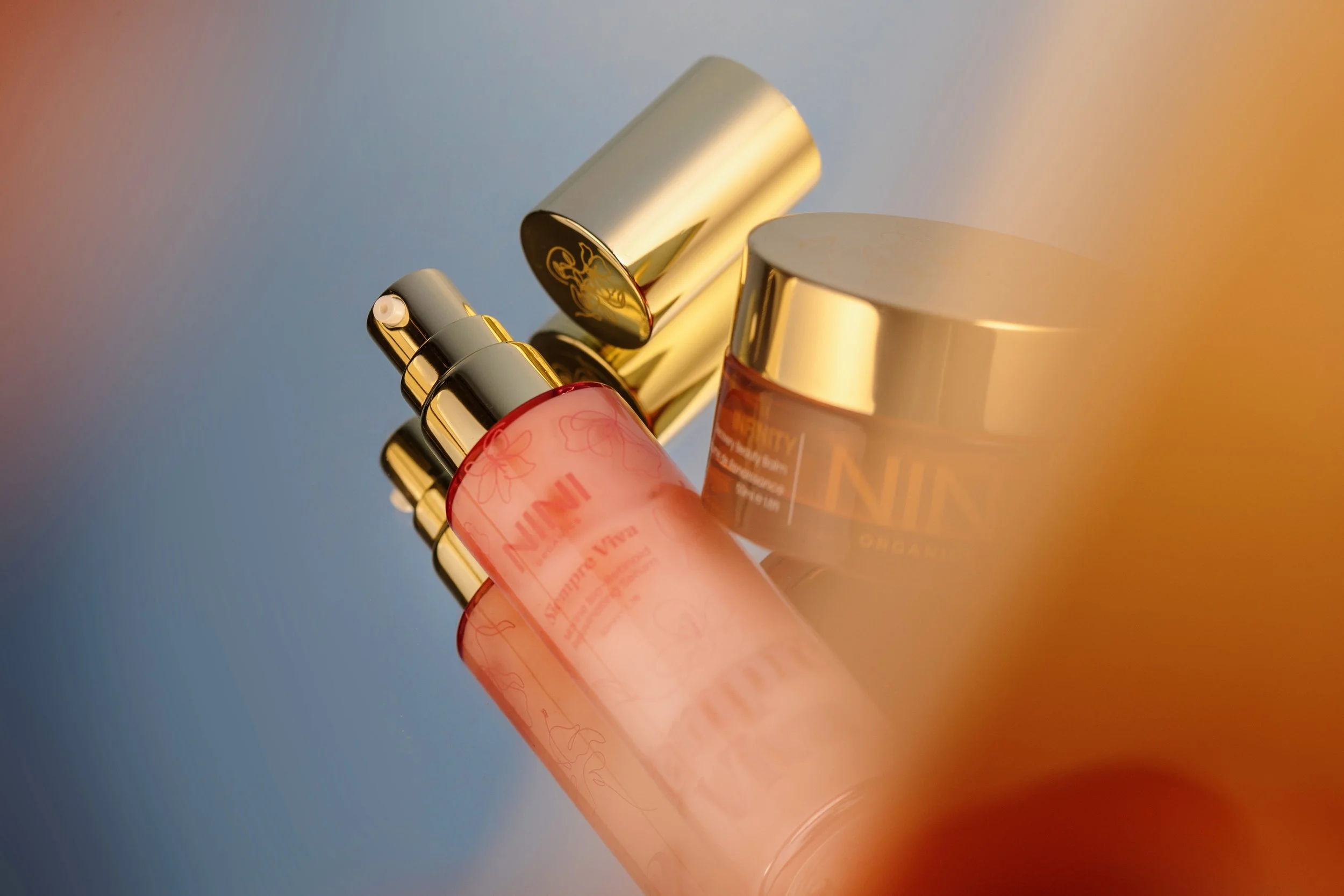 Close-up of several skincare or cosmetic bottles with metallic caps, set against a gradient background with warm and cool tones.