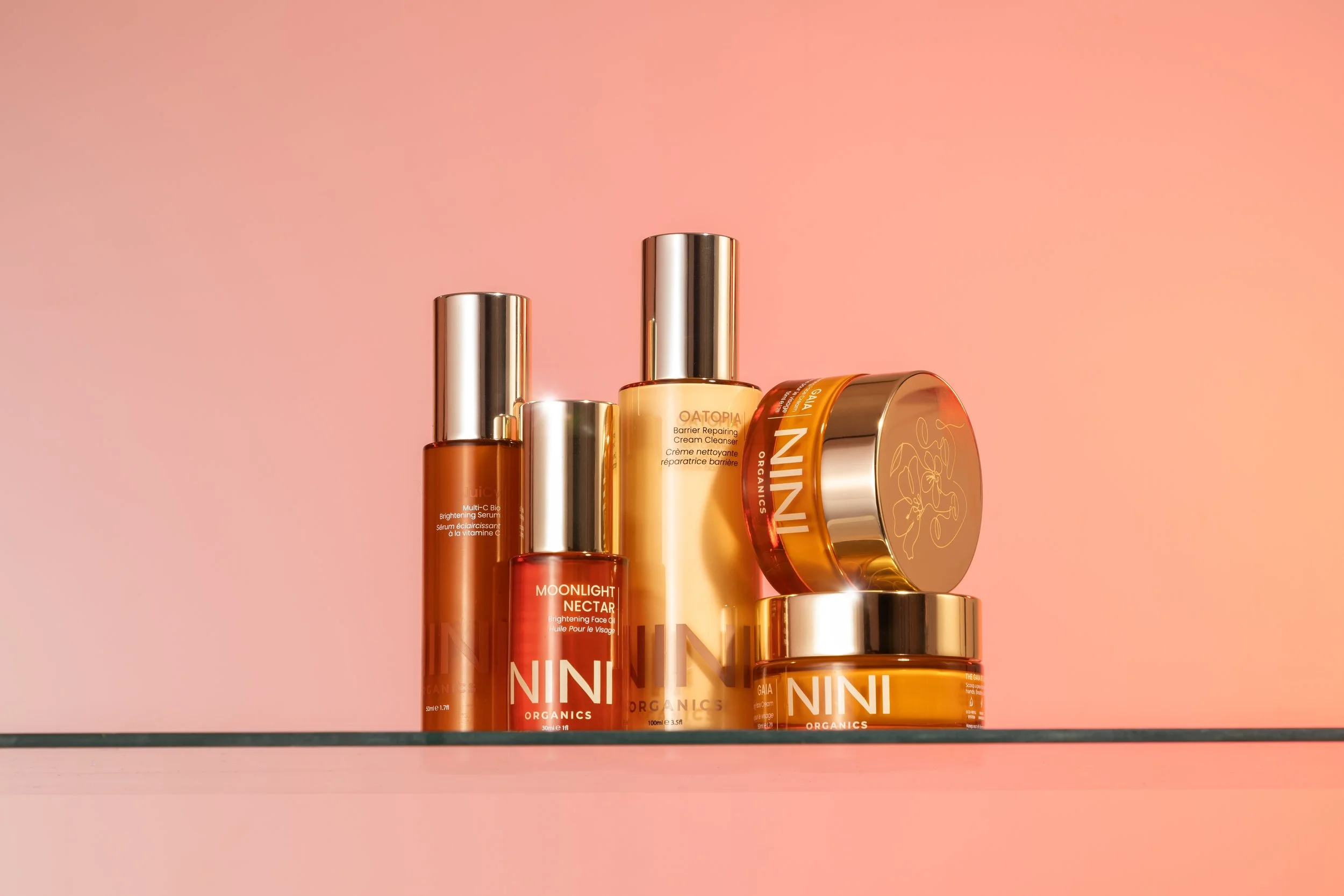 Various skincare products in orange, yellow, and red bottles and jars with silver caps on a glass shelf against a pink background.
