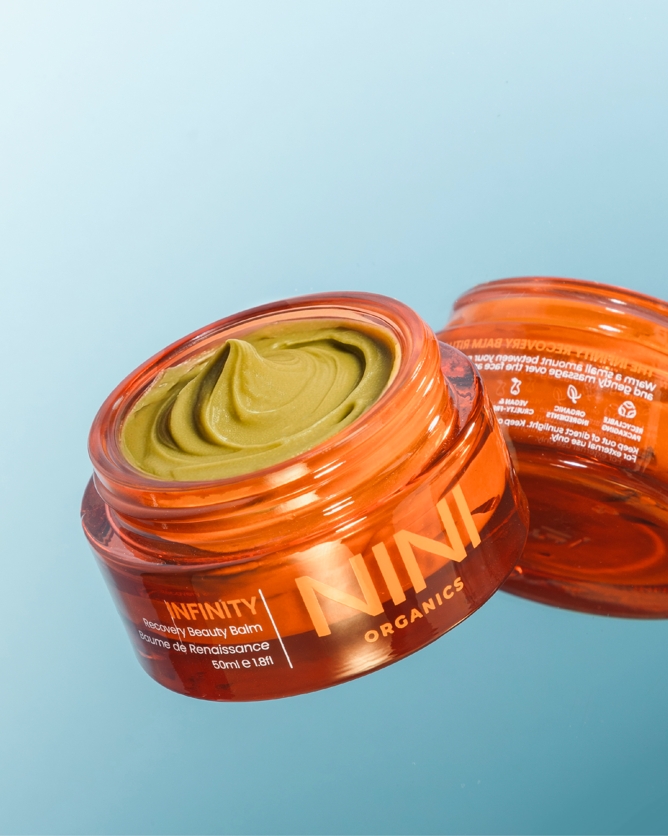 Close-up of an open jar of organic recovery beauty balm with greenish-yellow substance inside, against a light blue background.