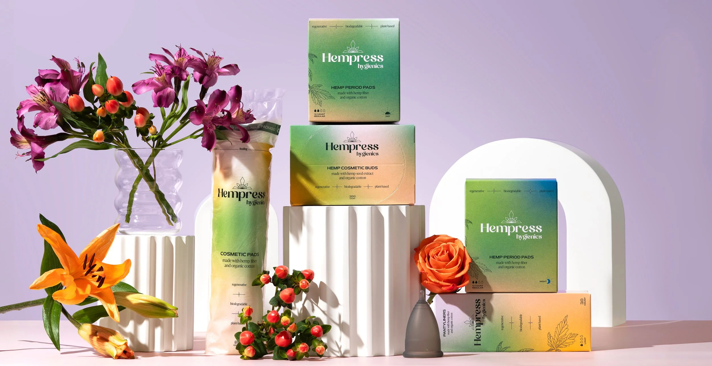 Various packages of Hempress hygienics products, including hemp period pads and cosmetic buds, arranged on white pedestals with flowers and a purple background.