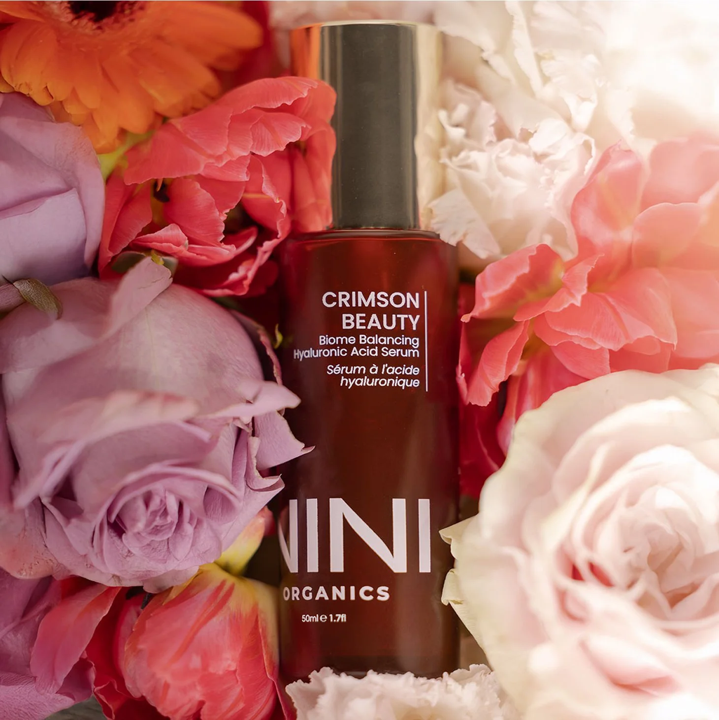 A bottle of Crimson Beauty Biome Balancing Hyaluronic Acid Serum surrounded by pink, peach, cream, and white roses.