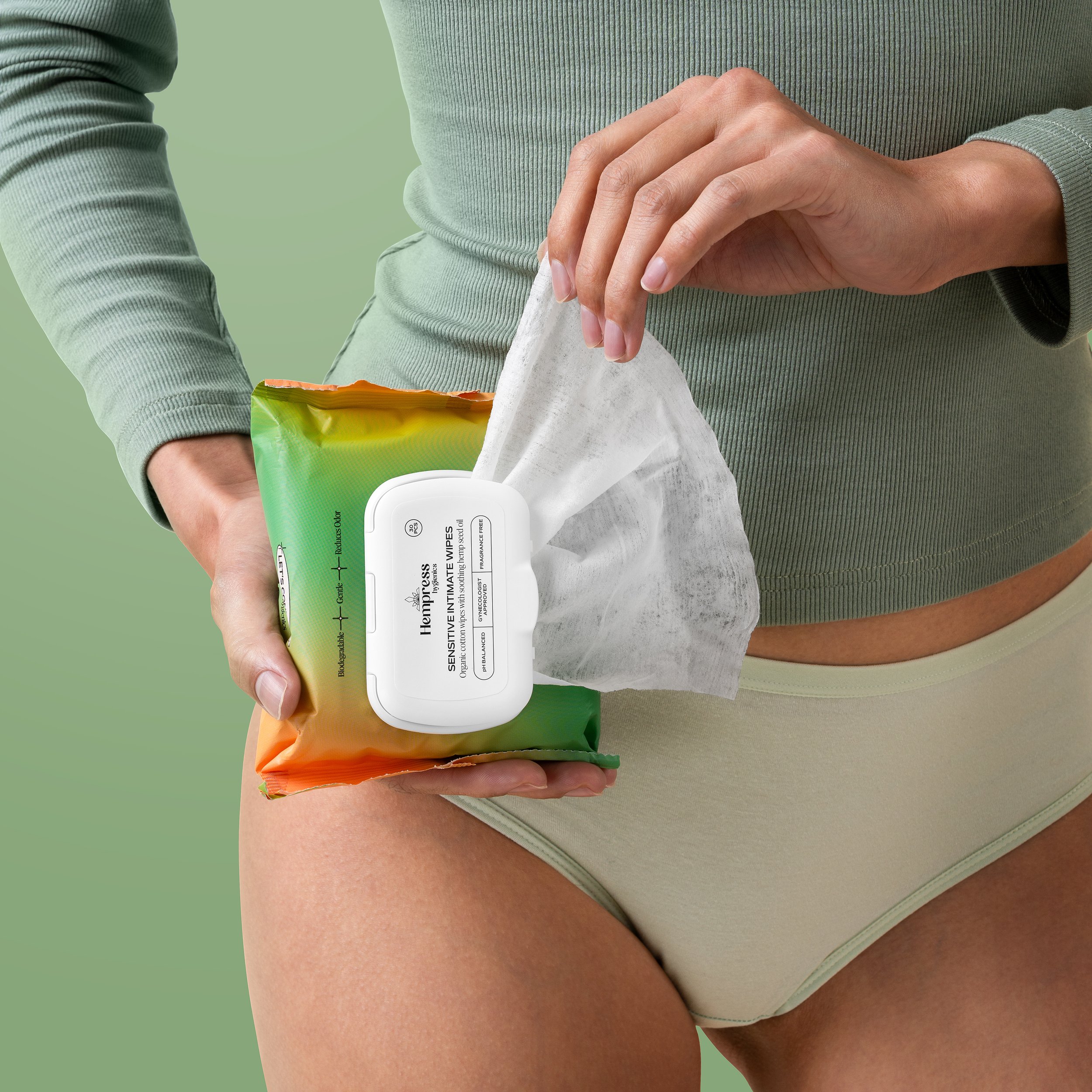 A person wearing a light green long sleeve shirt and light green shorts is holding a pack of wipes, with the top of the pack being green and orange, and a white container of wipes in the foreground.
