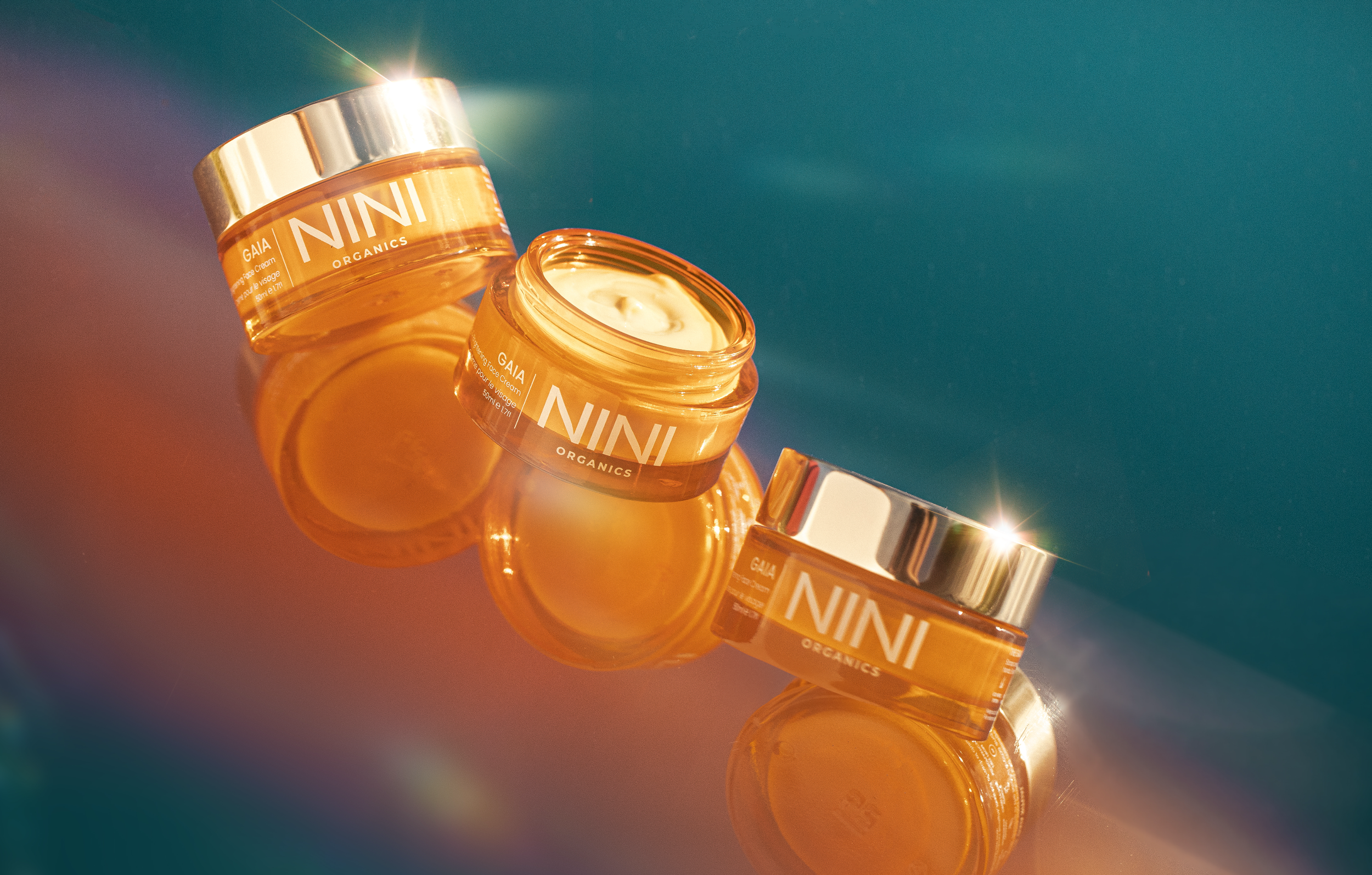 Multiple orange jars labeled 'NINI ORGANICS' with open cream in one jar, set against a vibrant teal background.