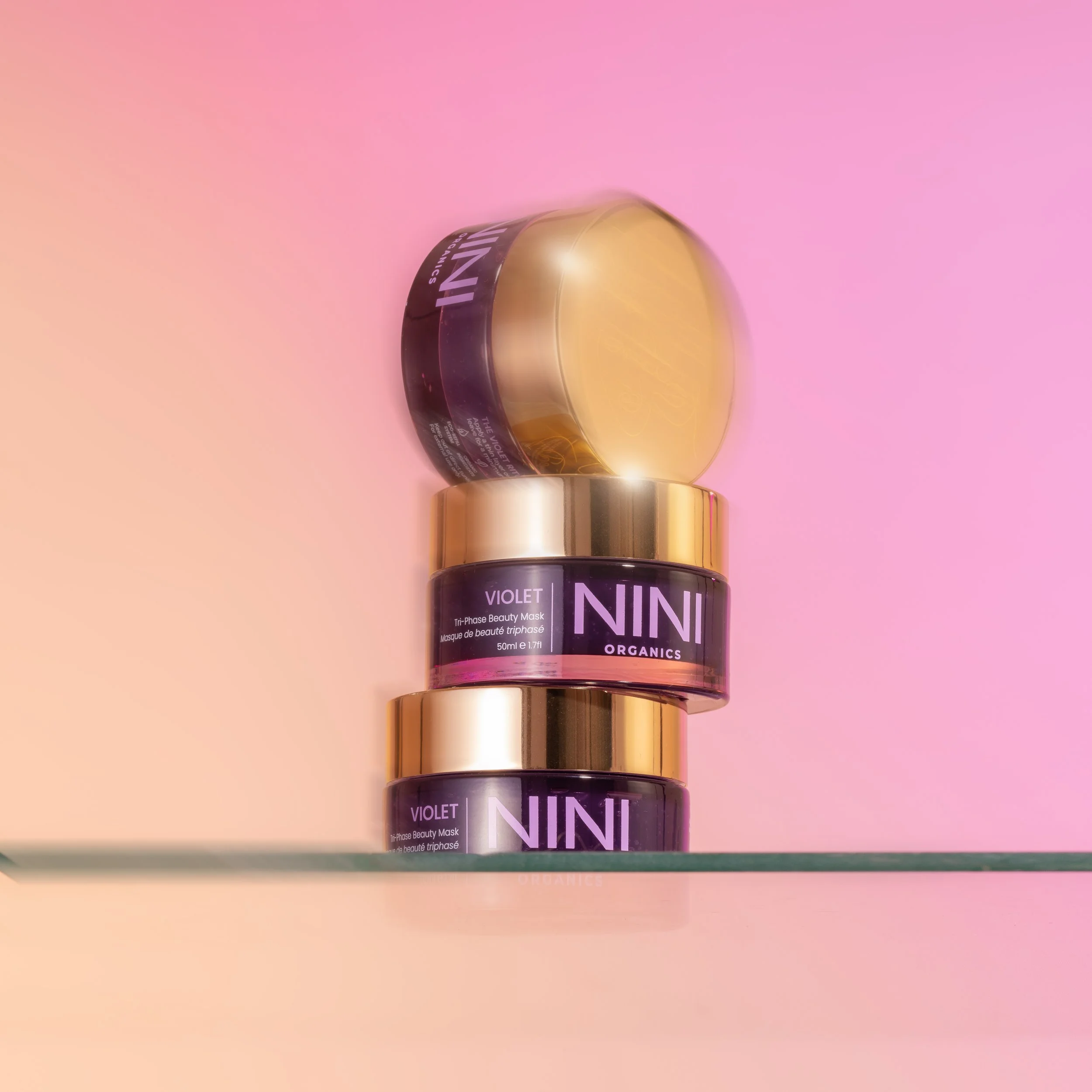 A small glass container with a black label reading "Violet Nini Organics" and a gold lid, placed on a pink background.