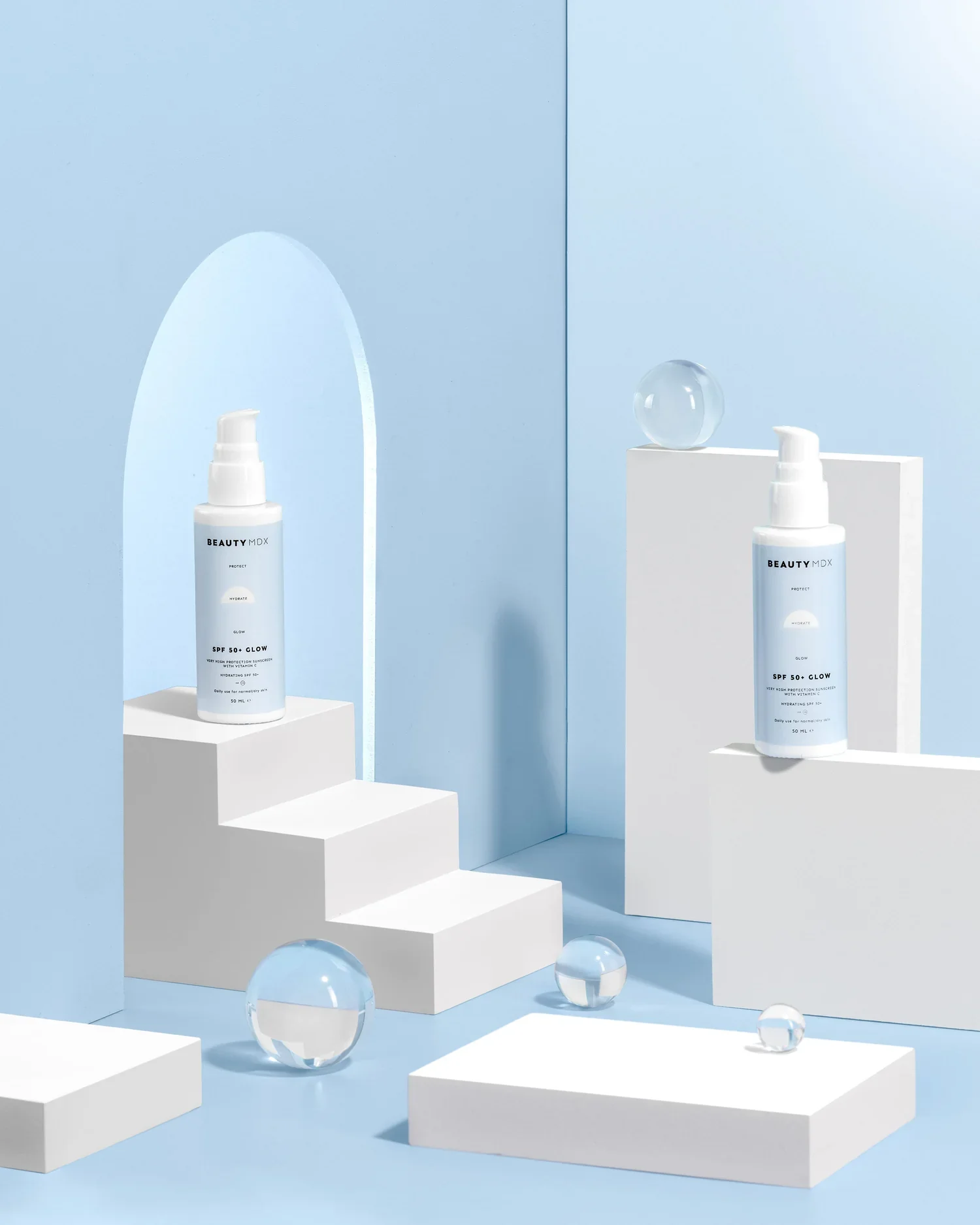 Two bottles of sunscreen spray labeled 'BEAUTY MDX SPF 50+ GLOW' are displayed on white geometric pedestals with glass spheres and a mirror, set against a blue background.