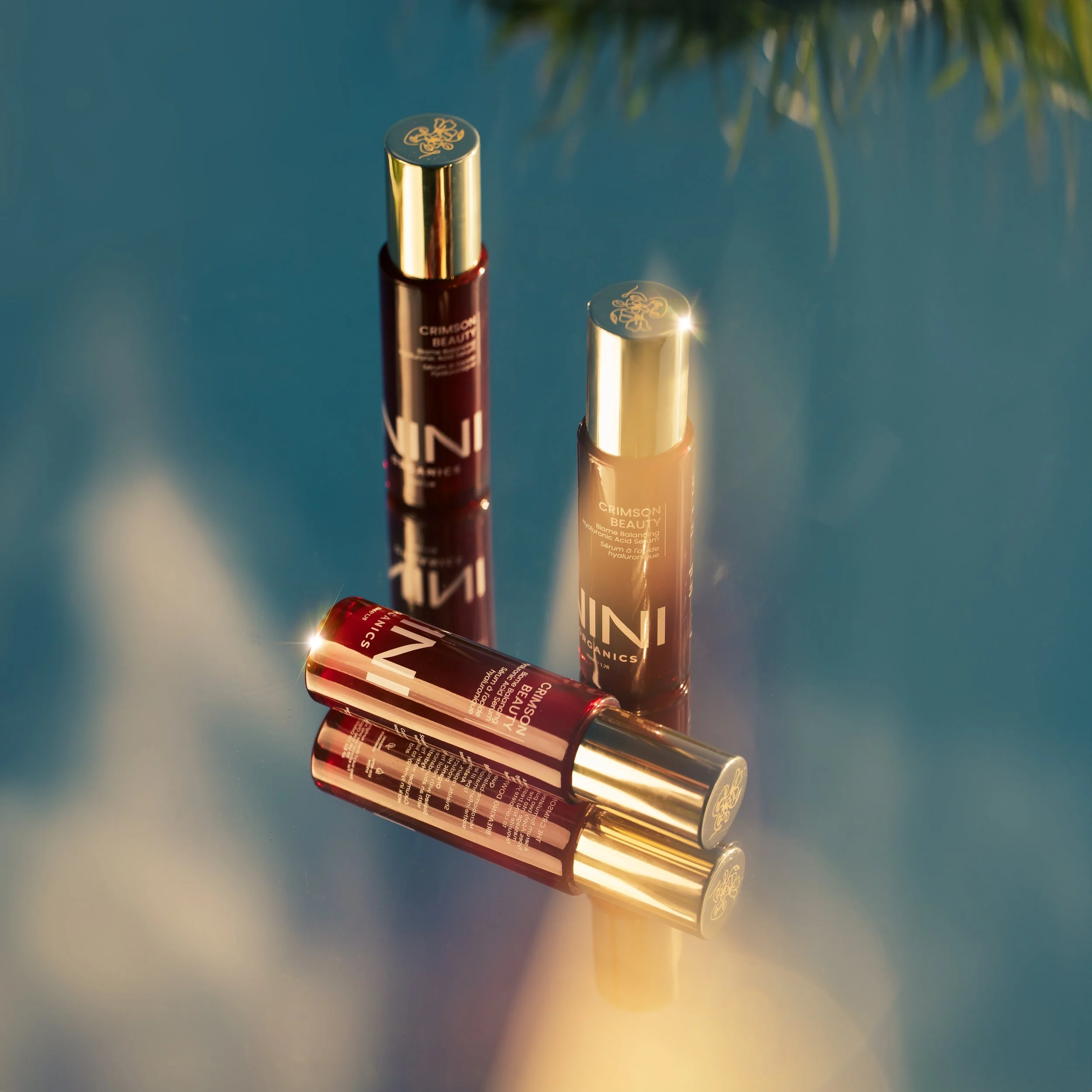 Three tubes of beauty serum and lip gloss with reflective gold caps are arranged in a row on a reflective surface, with a gradient background of blue, orange, and yellow.