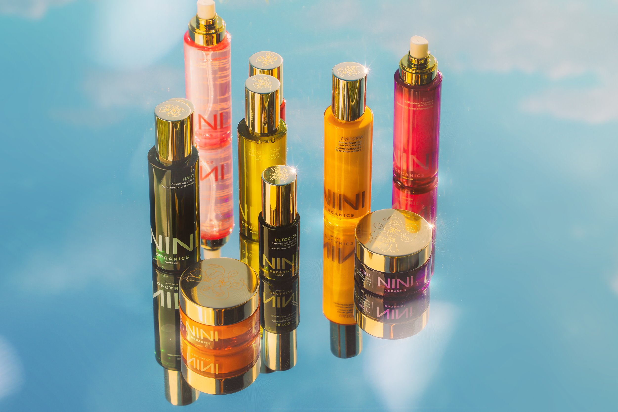 A collection of cylindrical skincare bottles and jars with metallic caps, arranged on a reflective blue surface. The bottles are in various colors including black, yellow, pink, and orange, with some displaying brand labels and product information. The arrangement creates reflections on the surface, with a bright light source causing highlights on the metallic caps.