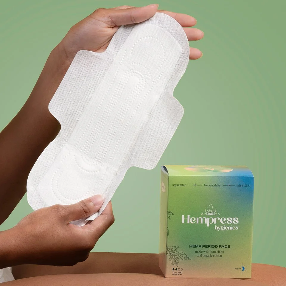 A person holding a large white detox or menstrual pad in their hand. A box of hemp period pads is on a table in front.