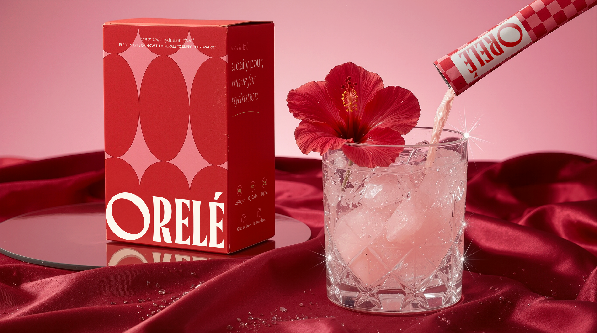 A pink box of Orélé electrolyte drink mix with a red hibiscus flower and pink drink in a crystal glass, set on a pink and red satin background.