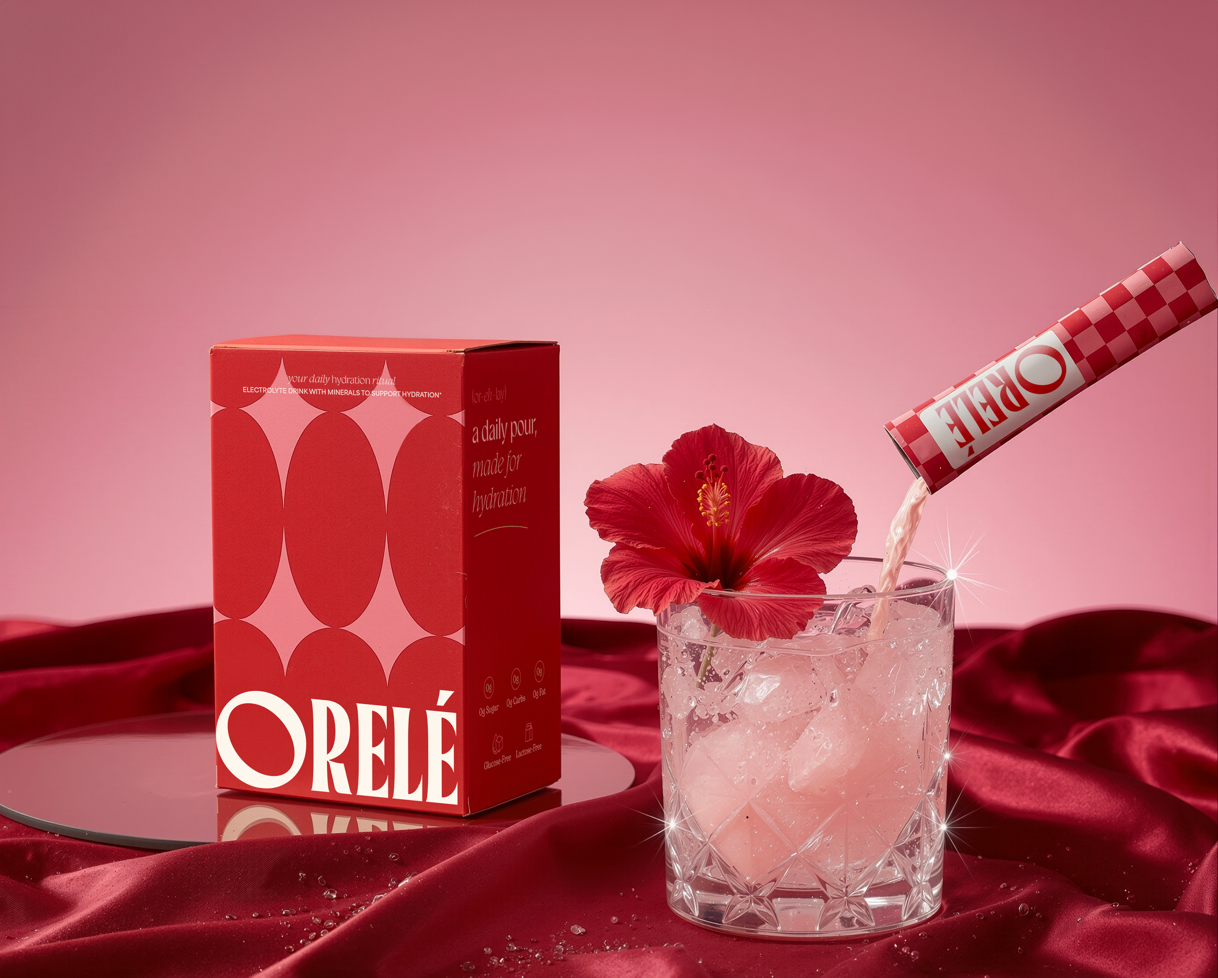 A carton of ORELEO hydration powder, a pink flower, and a glass of water with ice and pouring pink powder, set on a red velvet cloth with water droplets.