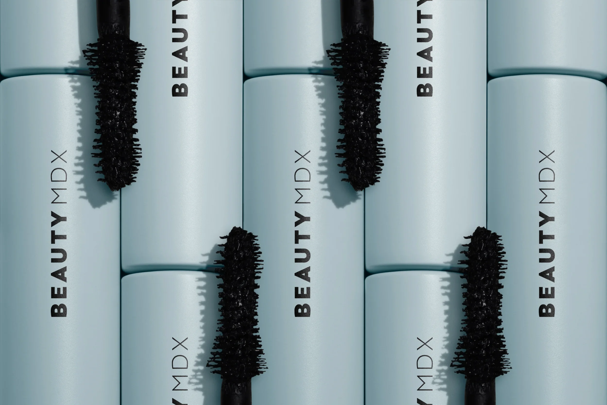 Multiple light blue cylindrical containers with black mascara wands resting on top, with the text 'BEAUTY MD X' printed on the containers.