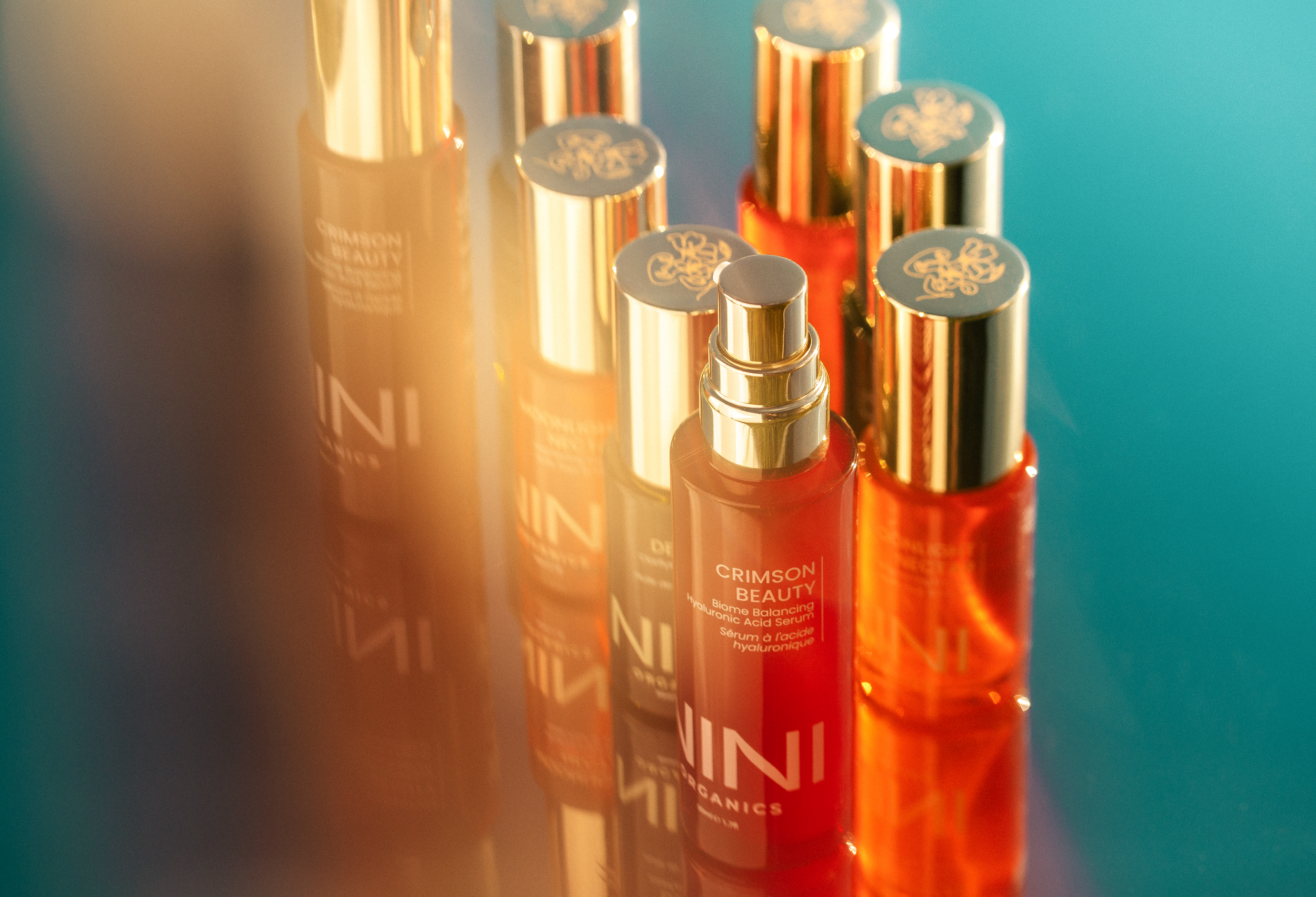 Close-up of orange skincare serum bottles with gold caps lined up, with focus on the front bottle labeled "Crimson Beauty" on a teal background.