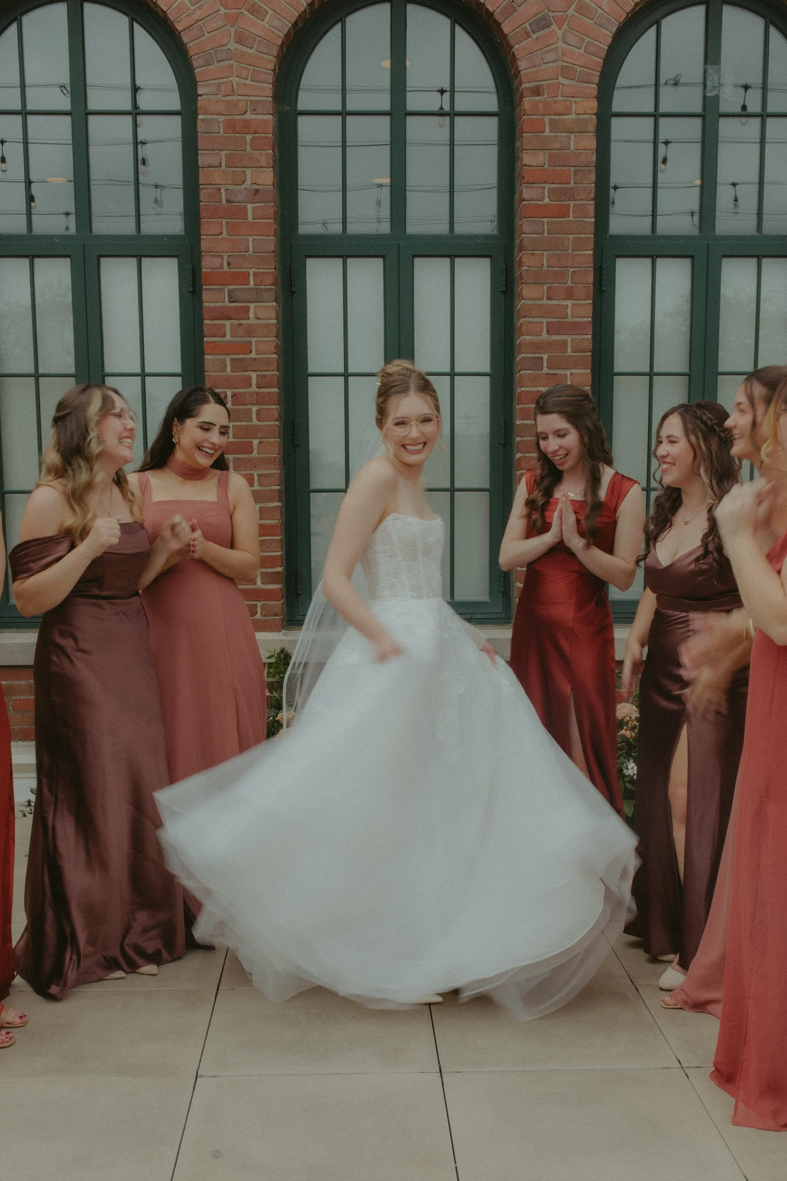 A bride with glasses in a white wedding dress standing among her bridesmaids in maroon and pink dresses, all smiling and joyfully interacting in front of a red brick wall with large green windowpanes.