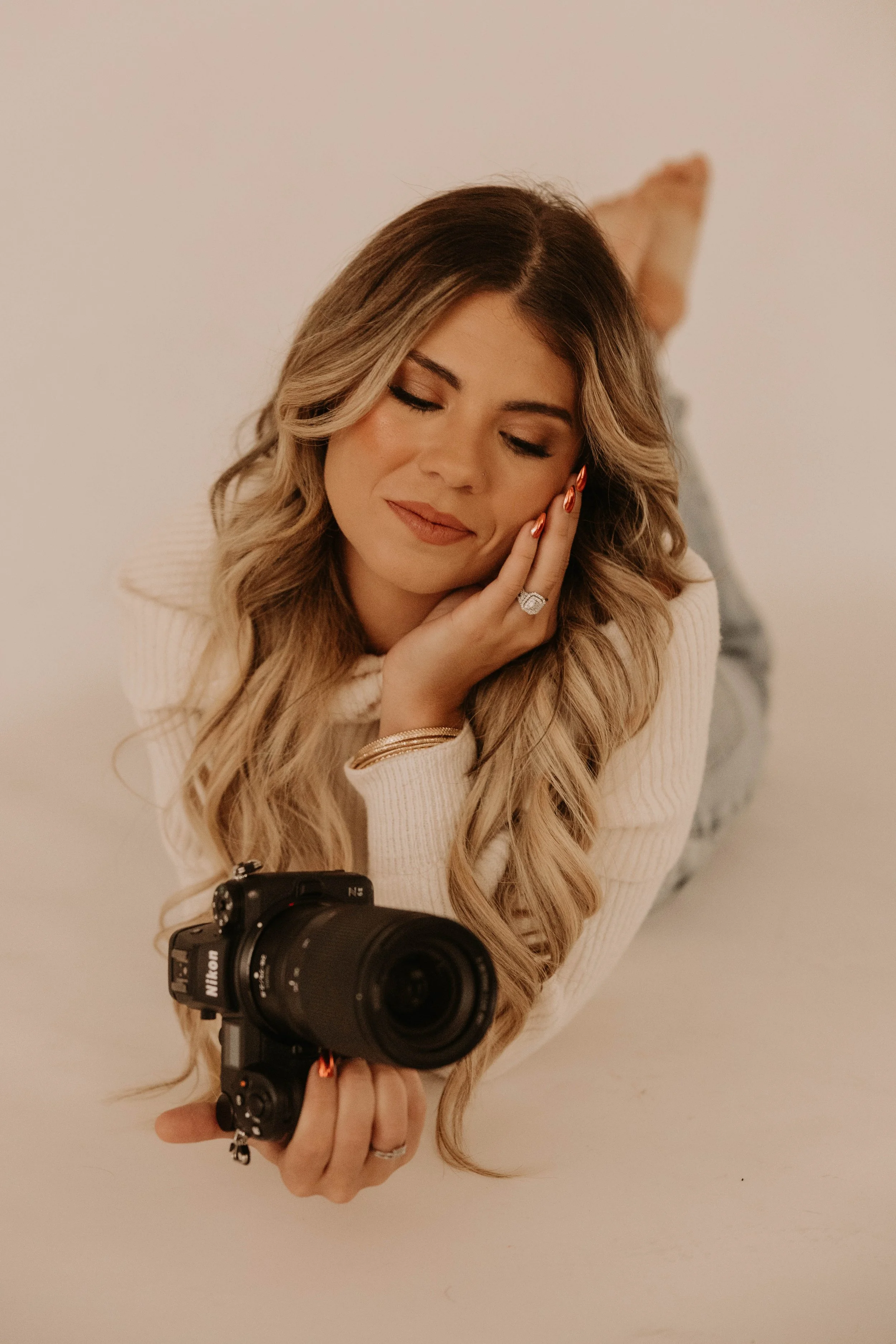 A woman with long wavy blonde hair taking a selfie with a camera. She is wearing a light-colored sweater and has her eyes closed, smiling softly. She rests her face on her hand, and her shoulder has a ring and bracelet. Her feet are up behind her, an