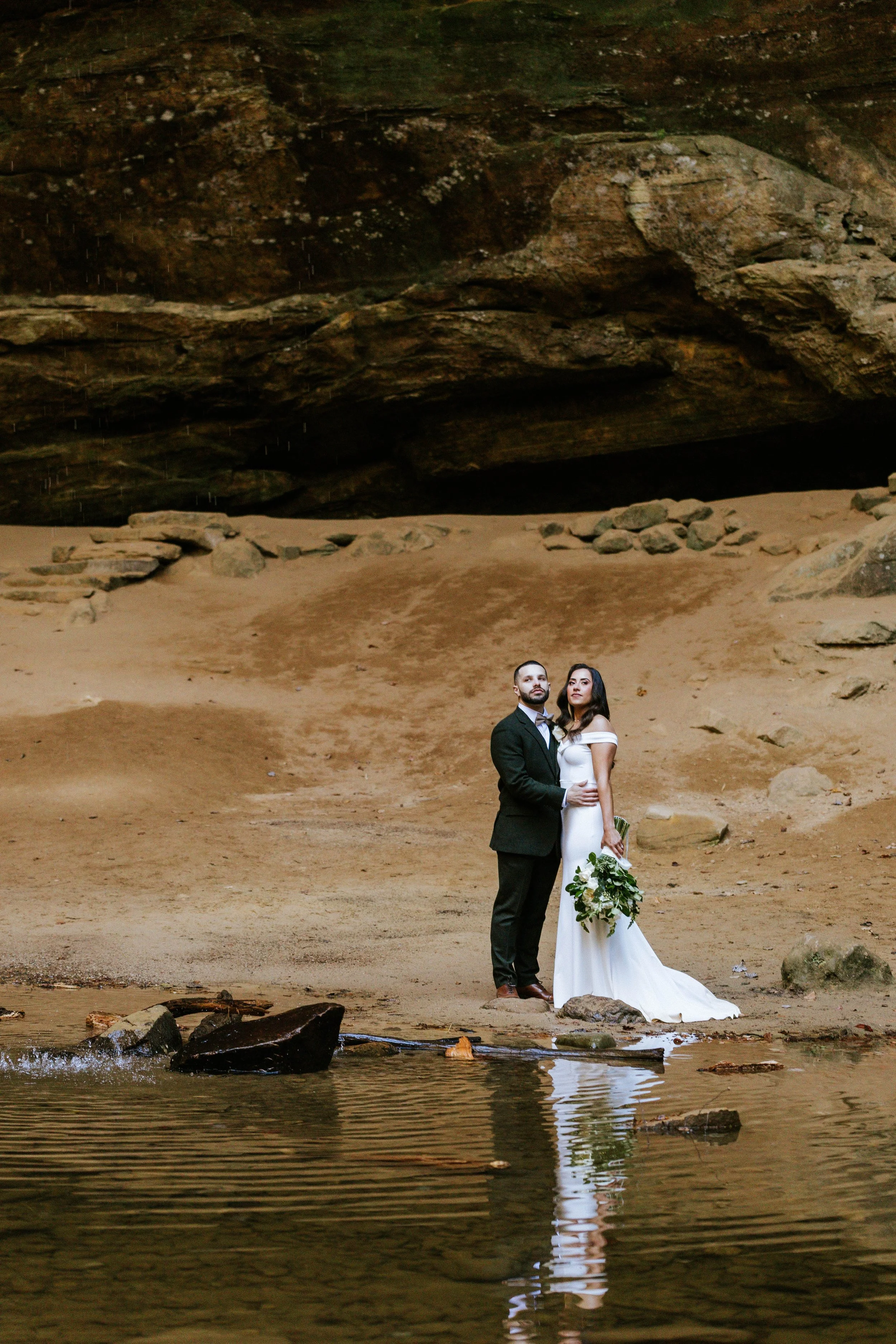 A bride and groom standing on a sandy beach near water with rocks, with a large rocky cliff behind them.
