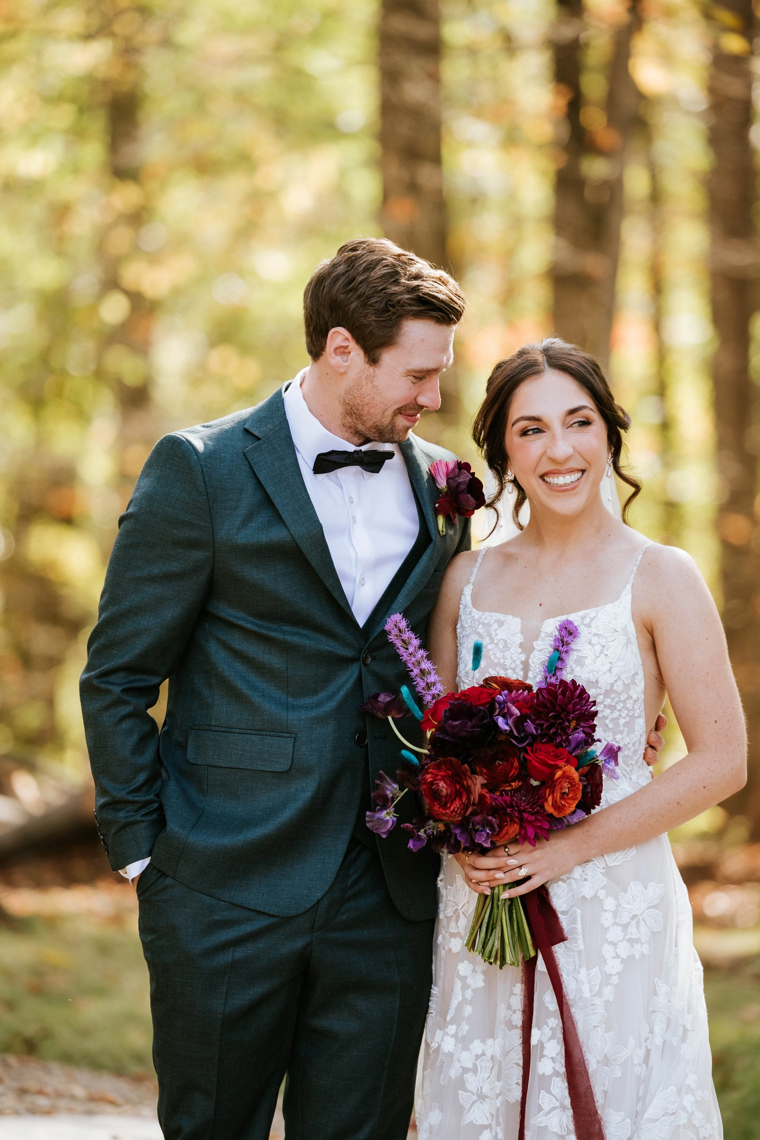 A smiling bride holding a bouquet of red, purple, and pink flowers, standing next to a groom in a dark suit with a black bow tie, in a forest setting during daytime.