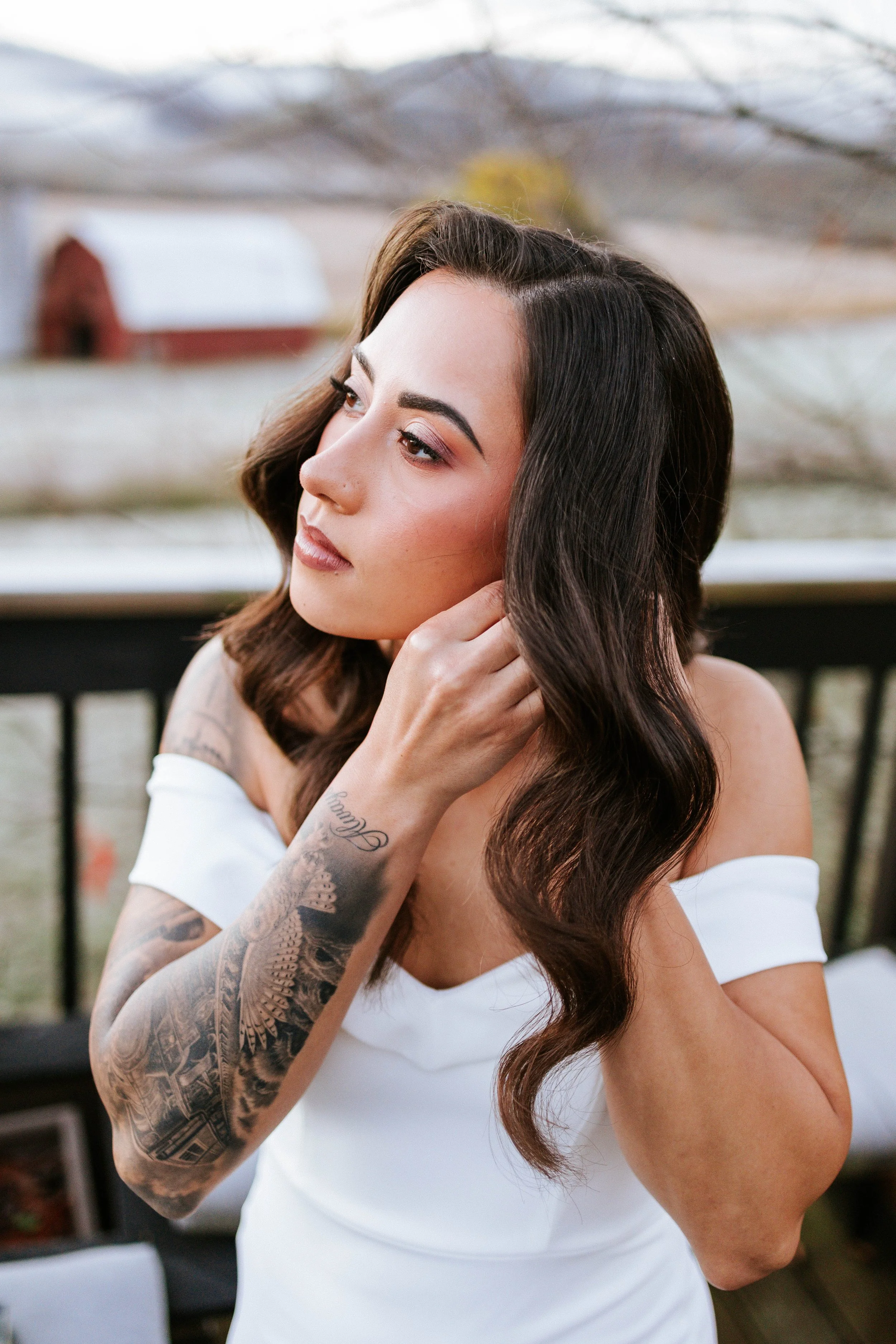 A young woman with long, dark, wavy hair and tattoos on her arm, wearing an off-shoulder white top, is adjusting her earring outdoors with a blurred background of trees and a building.