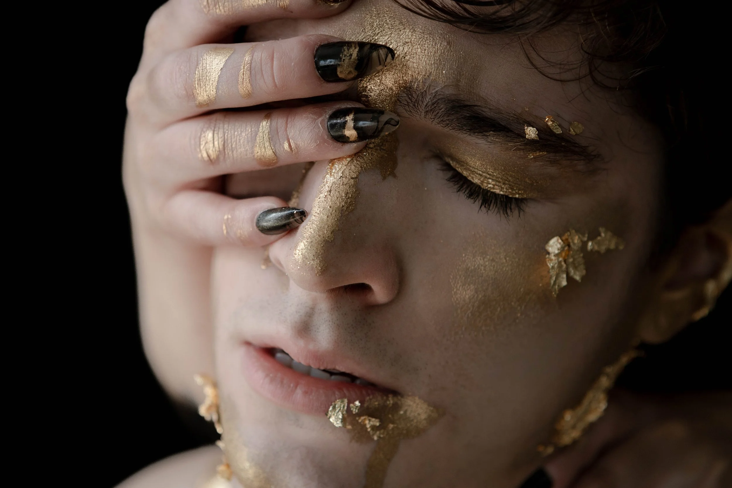 Close-up of a person's face with gold makeup and gold foil scattered on their face, with their hand covering part of their face and their eyes closed.