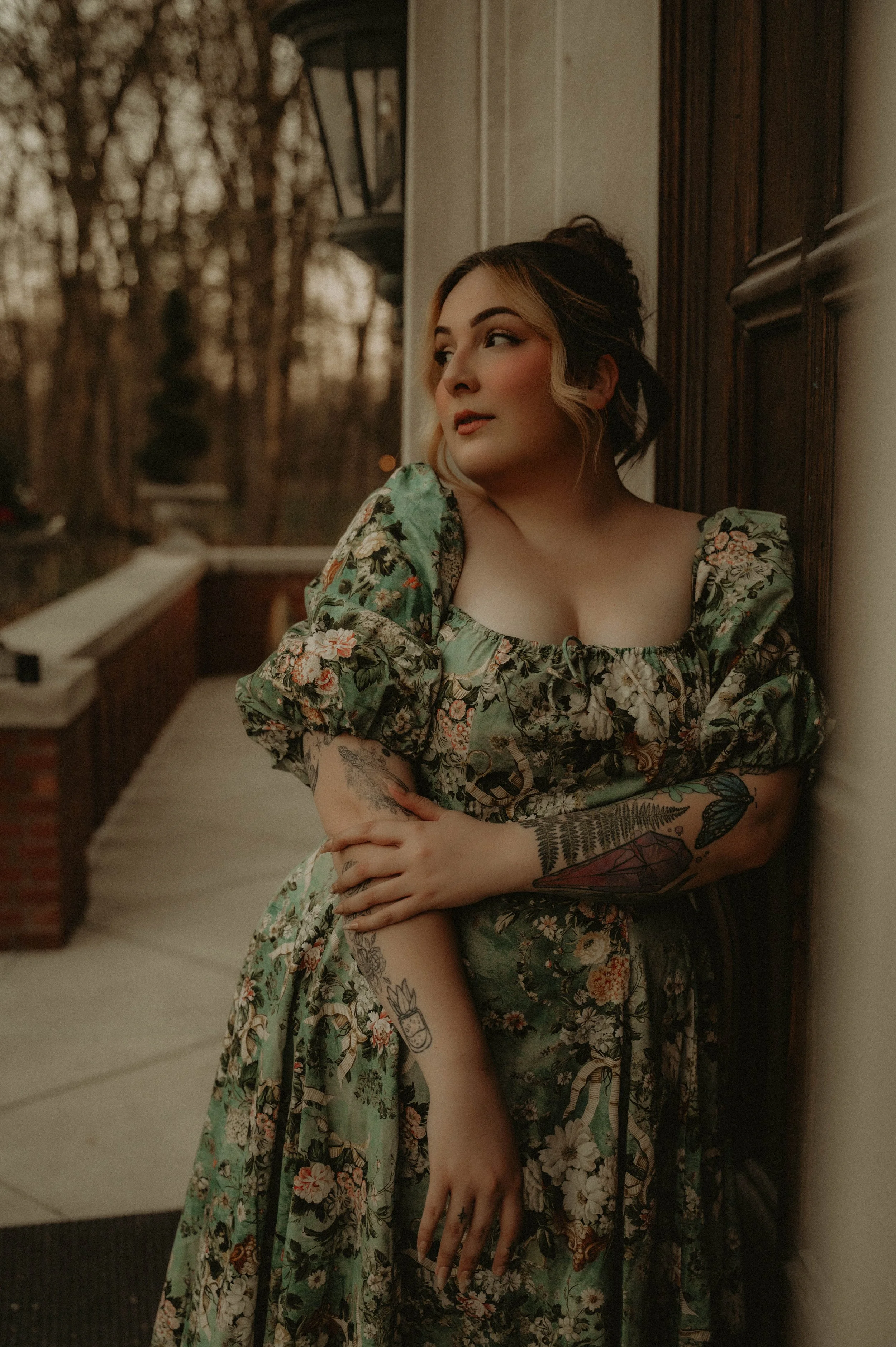 Woman with tattoos on her right arm and left thigh, wearing a green floral dress with puffy sleeves, standing against a dark wooden wall and looking to her left.