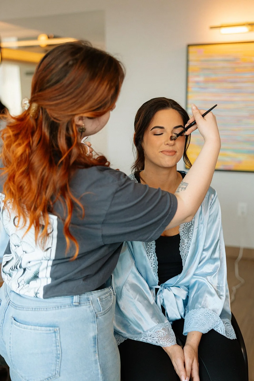 A makeup artist applying eyeshadow to a woman with closed eyes, seated indoors with colorful abstract wall art in the background.