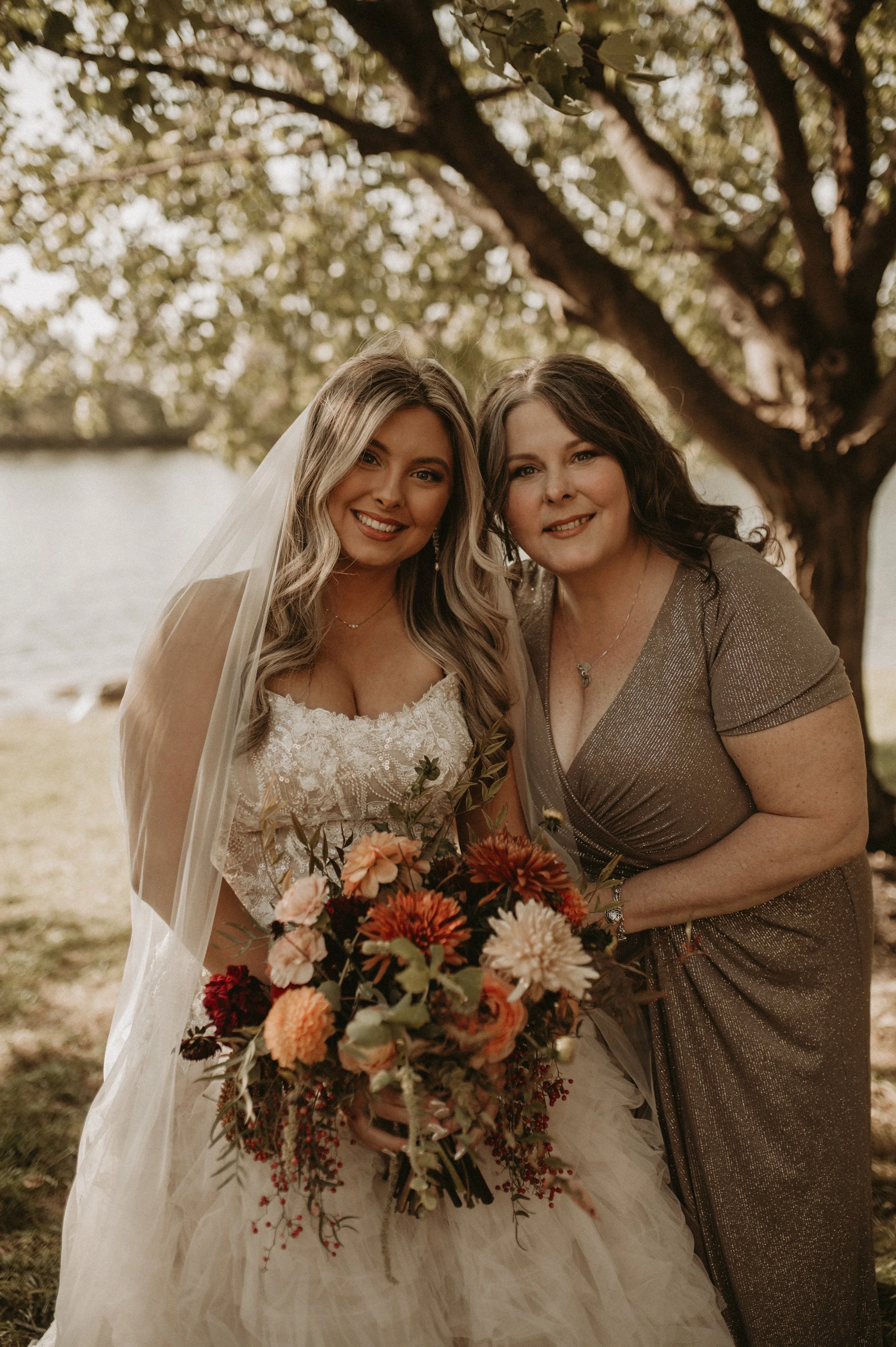 Two women standing near a tree outdoors, one wearing a wedding dress and veil, holding a bouquet of flowers, the other in a shimmery dress, smiling together.