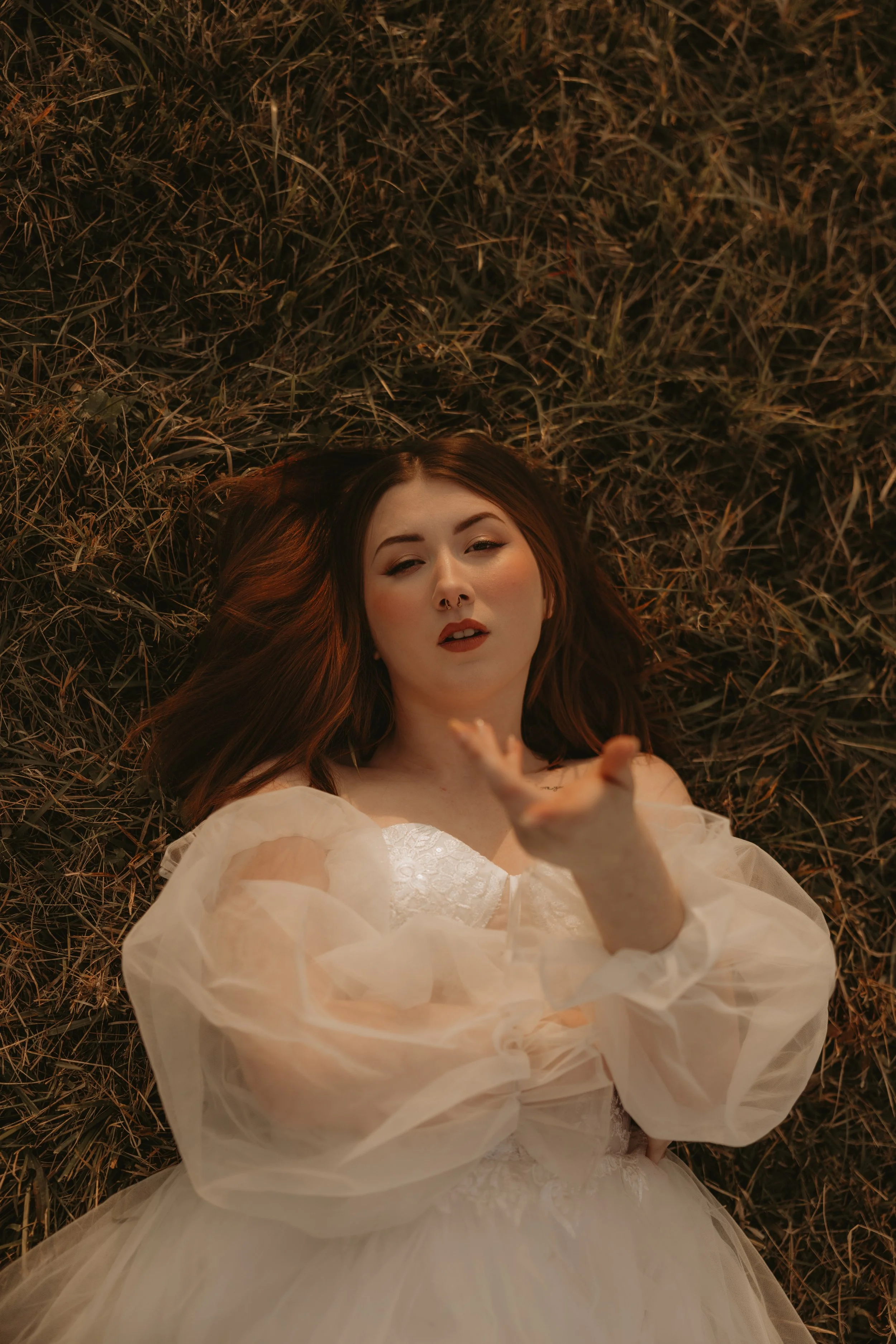 A woman with long red hair lying on dry grass, wearing a white dress with sheer sleeves, reaching out with her right hand.