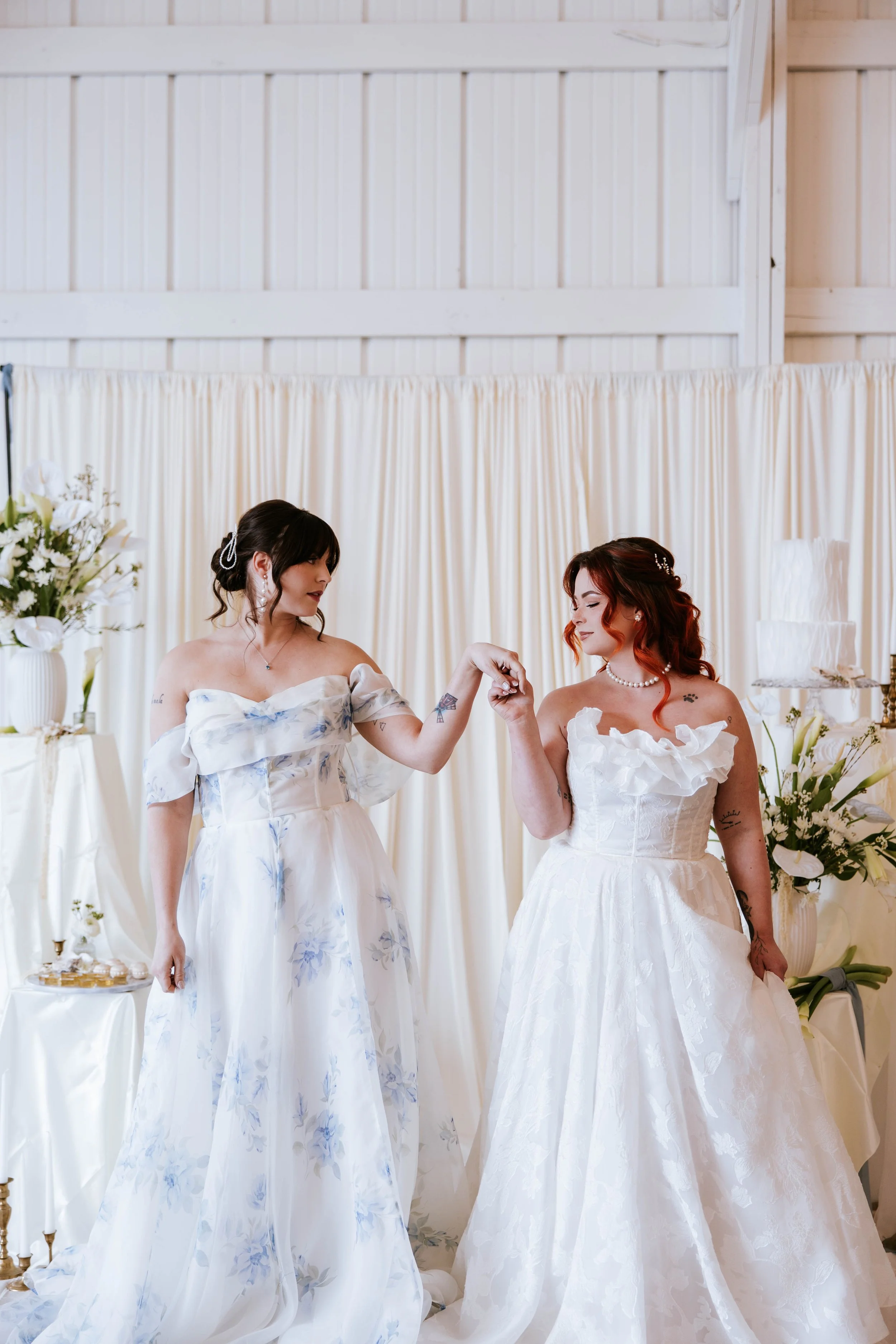Two women in wedding dresses holding hands at a wedding reception with white flowers and drapery background.