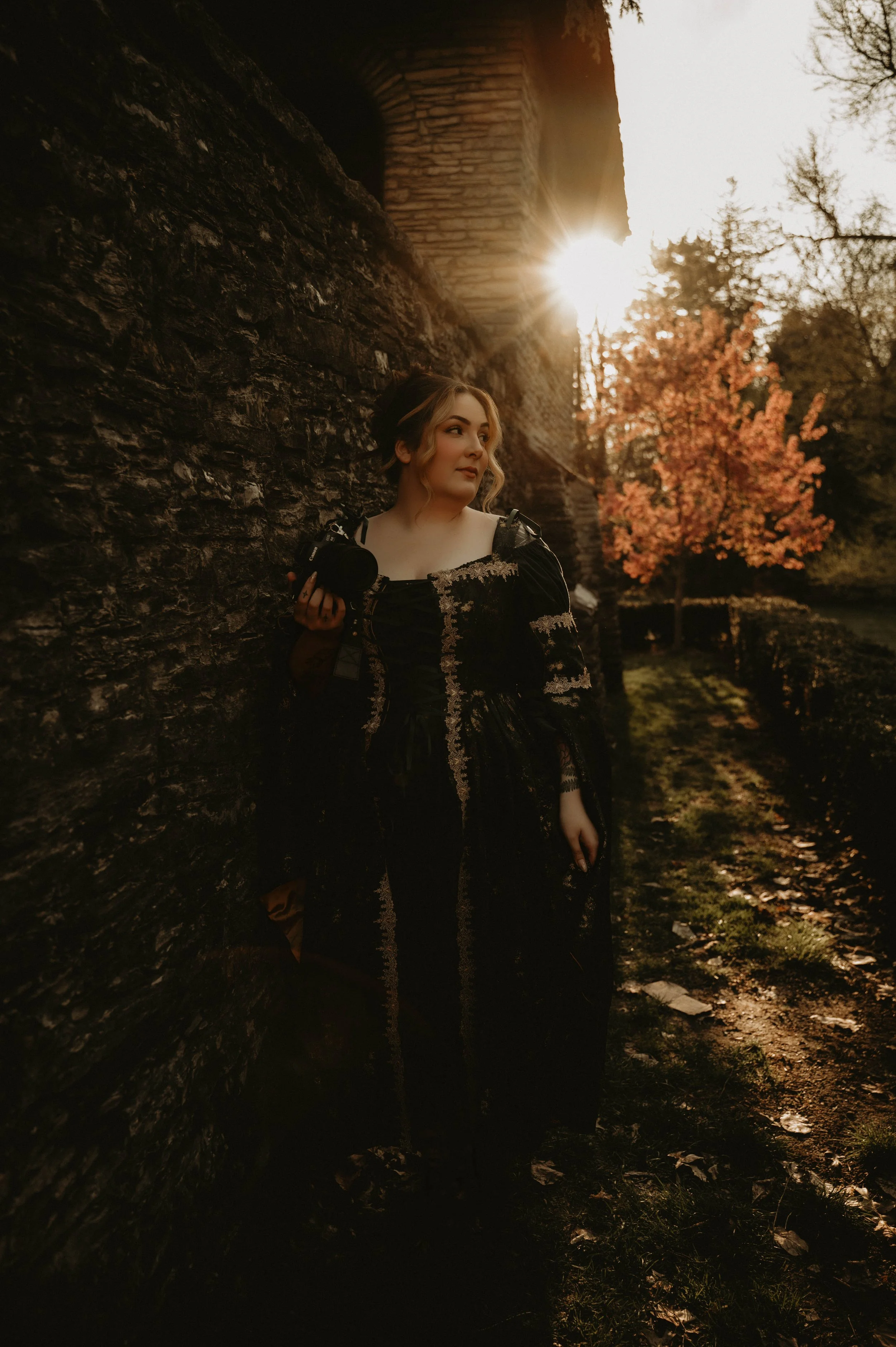 A woman with wavy blonde hair holding a camera, standing against a stone wall in an outdoor setting during sunset, with a tree with orange leaves in the background.