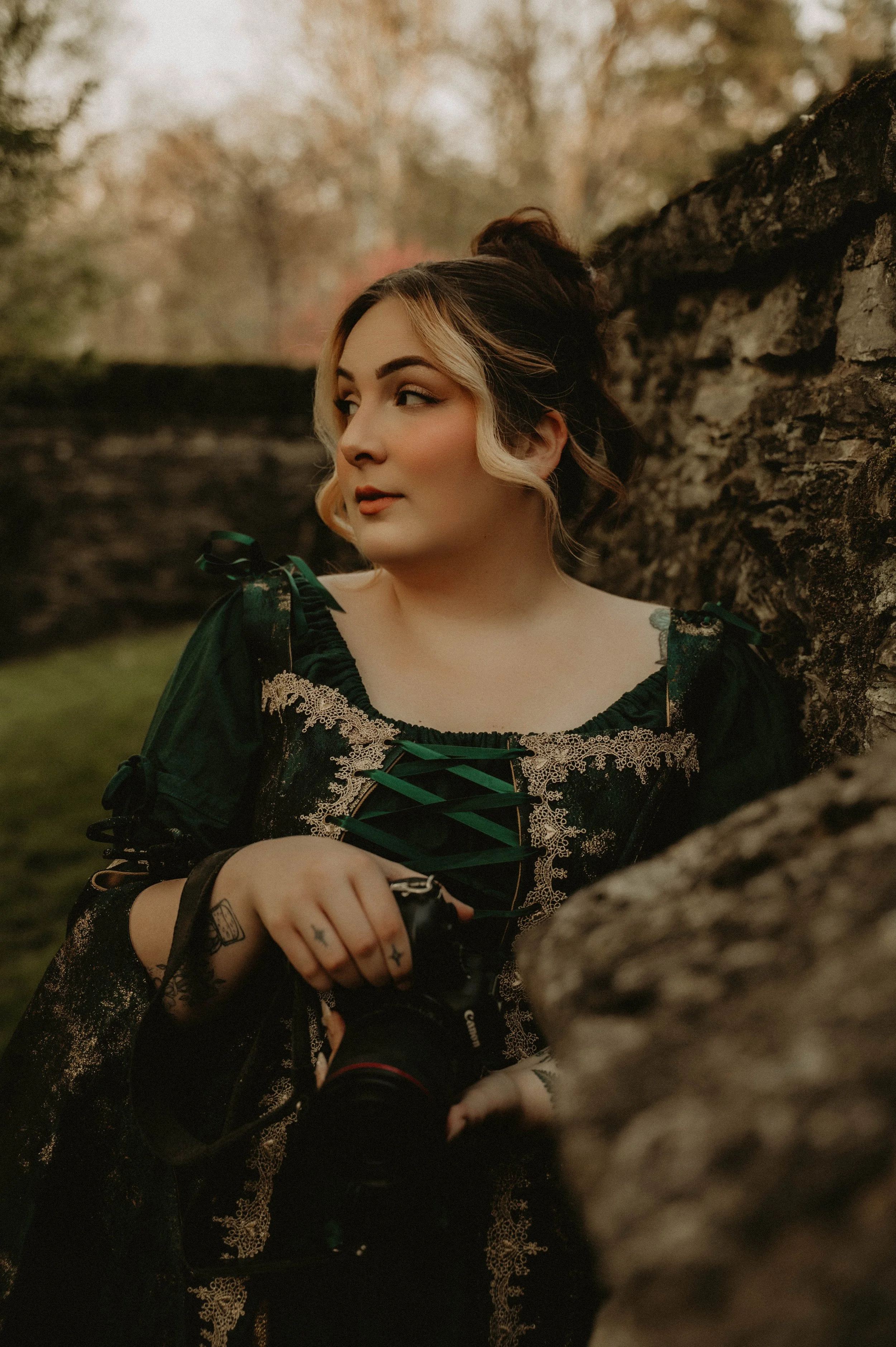 A woman with light skin and blonde hair styled in loose curls, wearing a dark green dress with lace and ribbon details, is sitting outdoors next to a stone wall or tree trunk. The background features blurred trees with fall foliage.