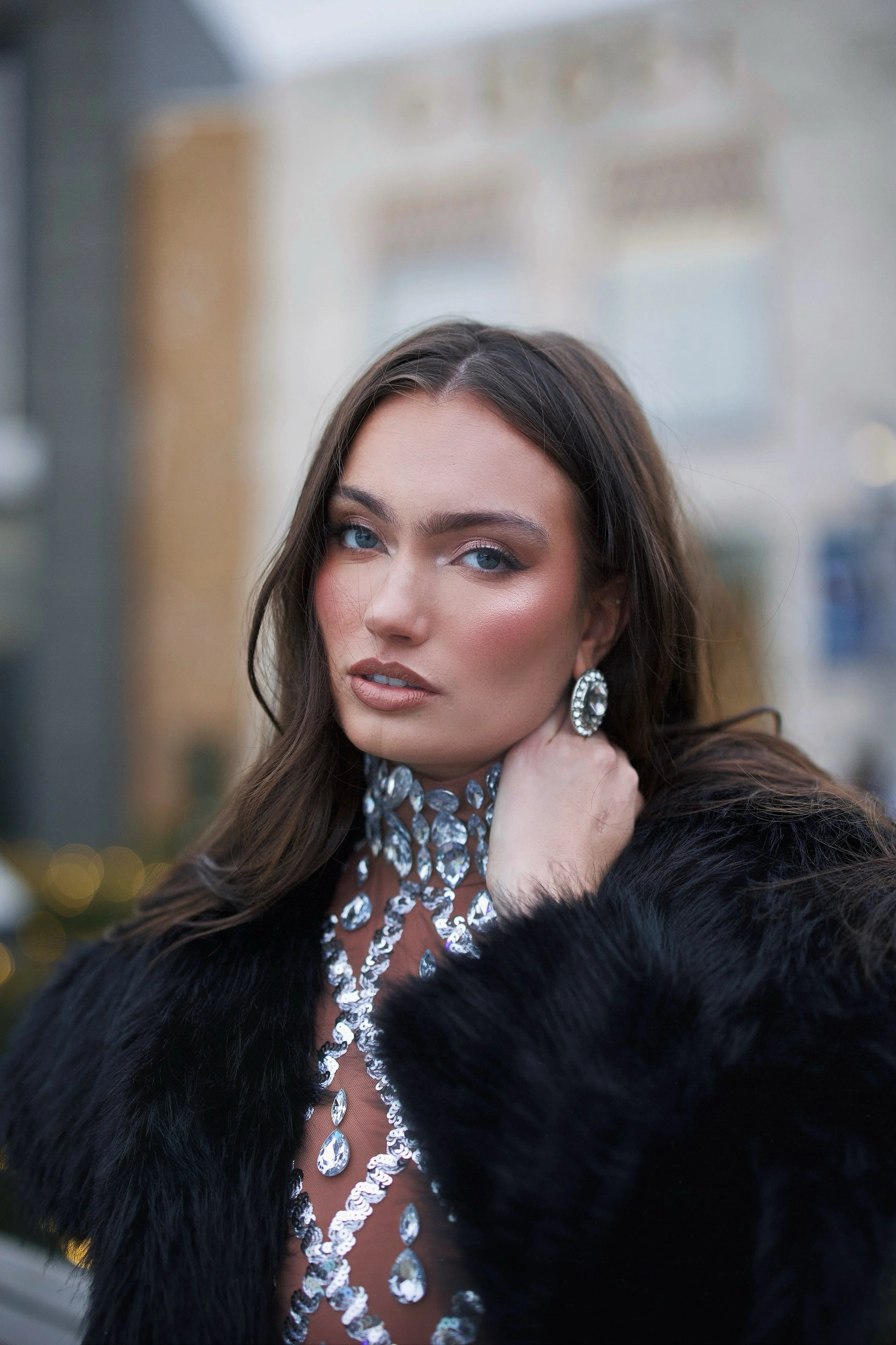 Close-up of a woman with long brown hair, blue eyes, makeup, wearing large diamond earrings and a diamond jewelry necklace, dressed in a black fur coat, in an outdoor setting with blurred background.