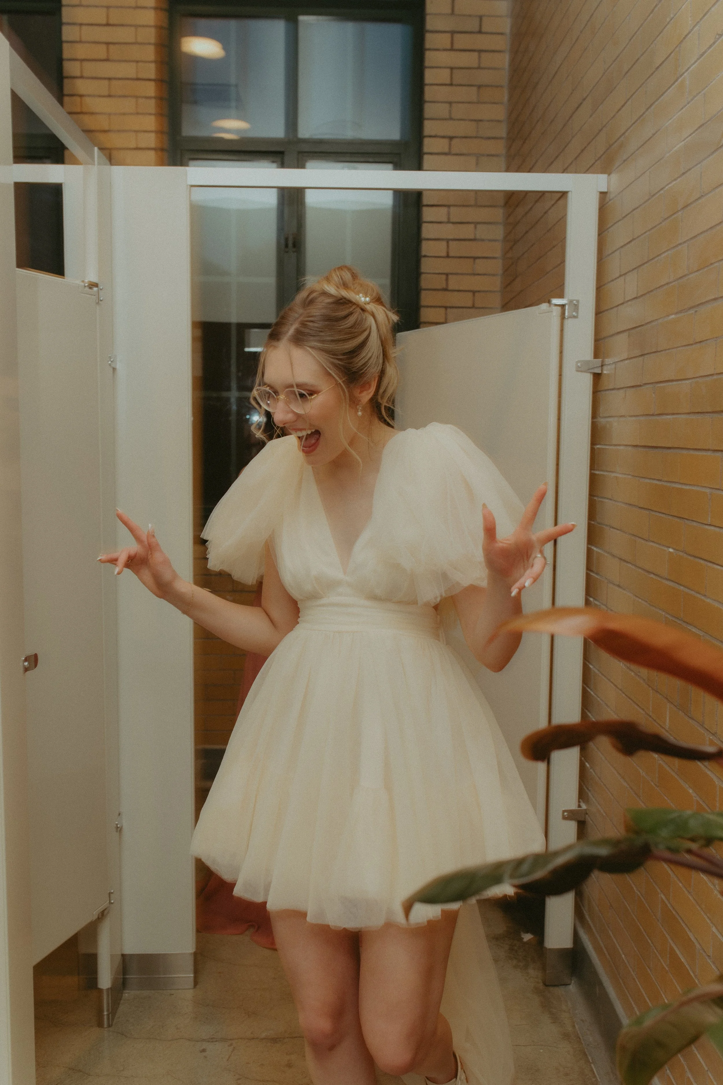 A woman in a cream-colored dress with tulle sleeves, glasses, and pearl earrings, joyfully posing with peace signs in a locker room with brick walls.