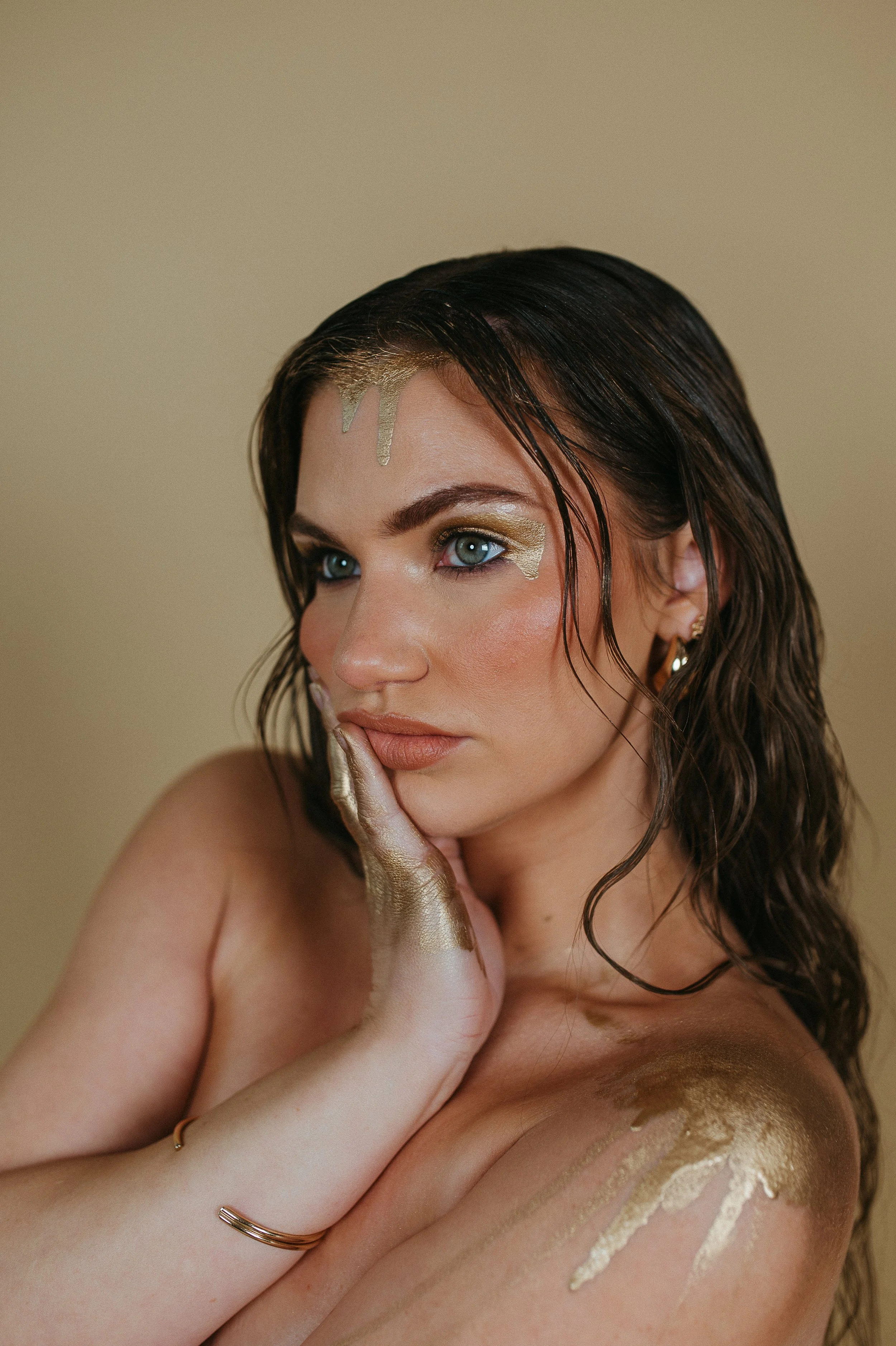 A woman with wet dark hair and blue eyes, decorated with gold paint or makeup on her face and body, looking contemplative with her hand near her face.