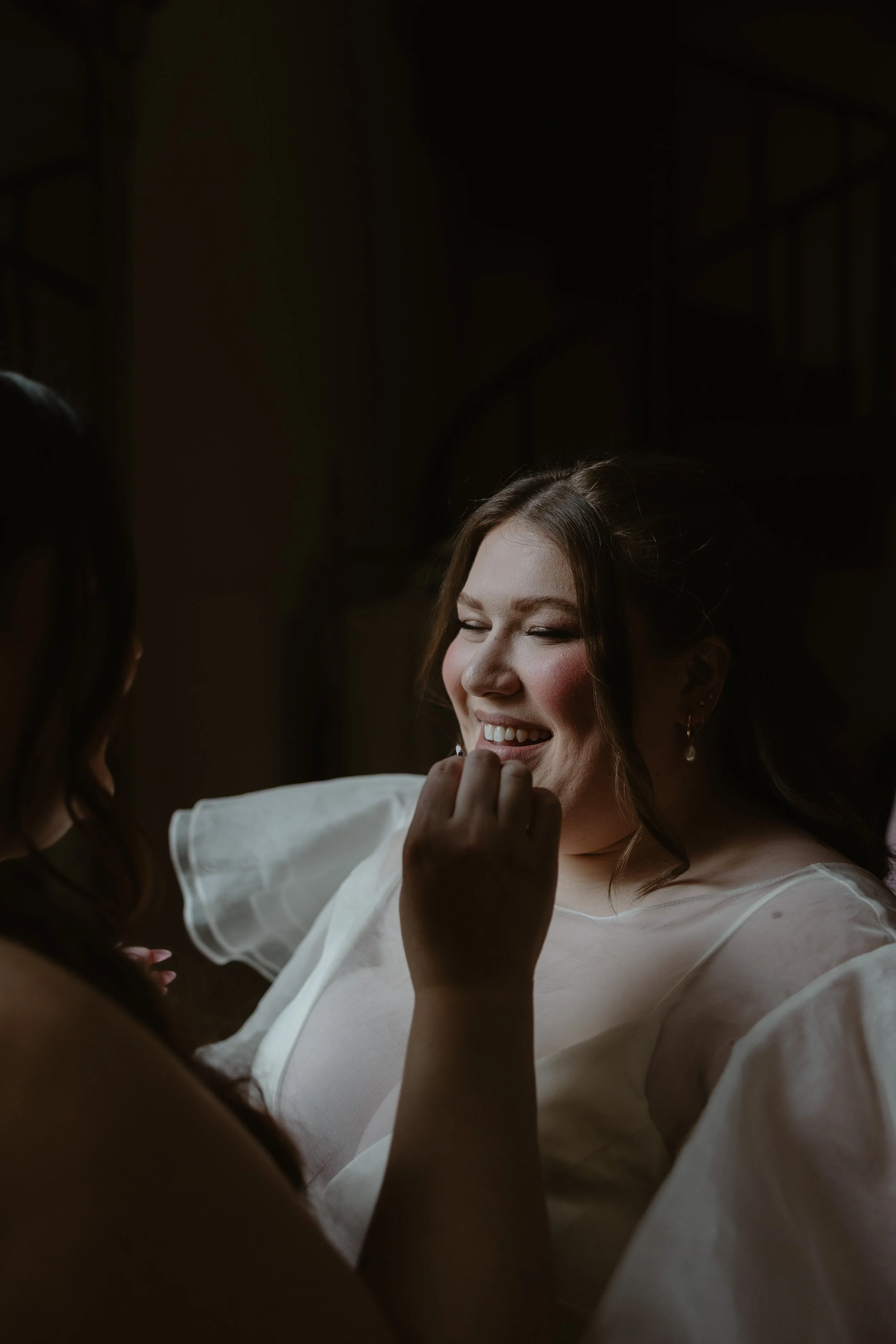 A woman with a radiant smile and closed eyes, wearing a white dress, is being assisted with makeup or a similar preparation by another woman in a dimly lit setting.