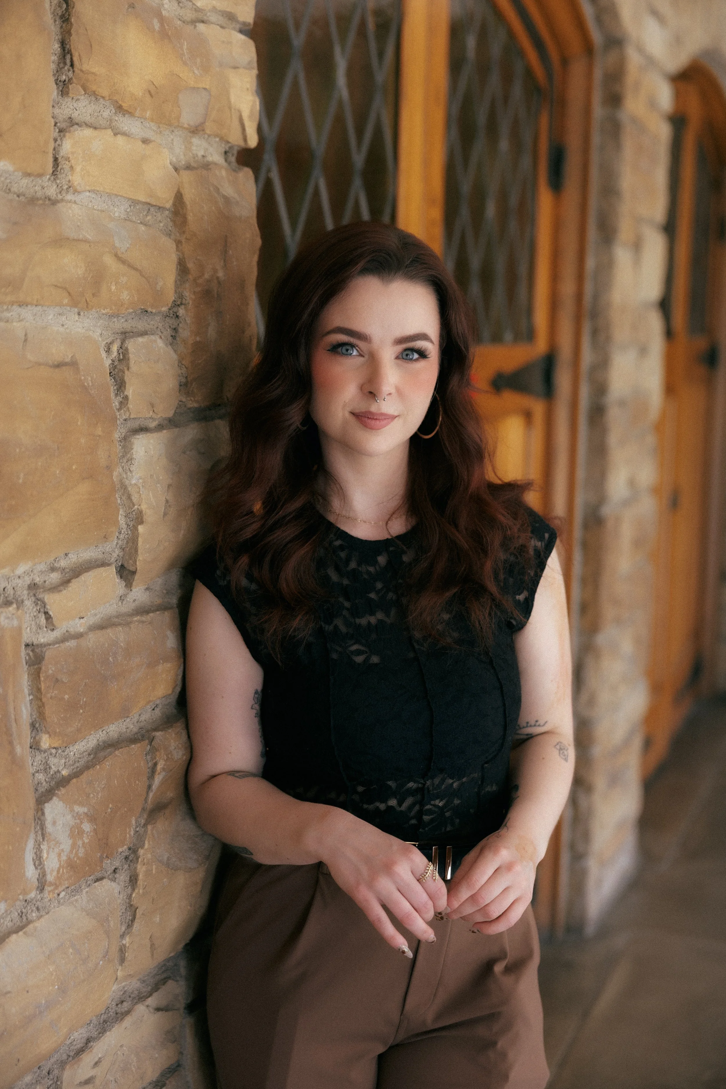 A young woman with dark red hair, blue eyes, and tattoos on her arms, poses against a rustic stone wall with wooden doors in the background. She is wearing a black top and brown pants.