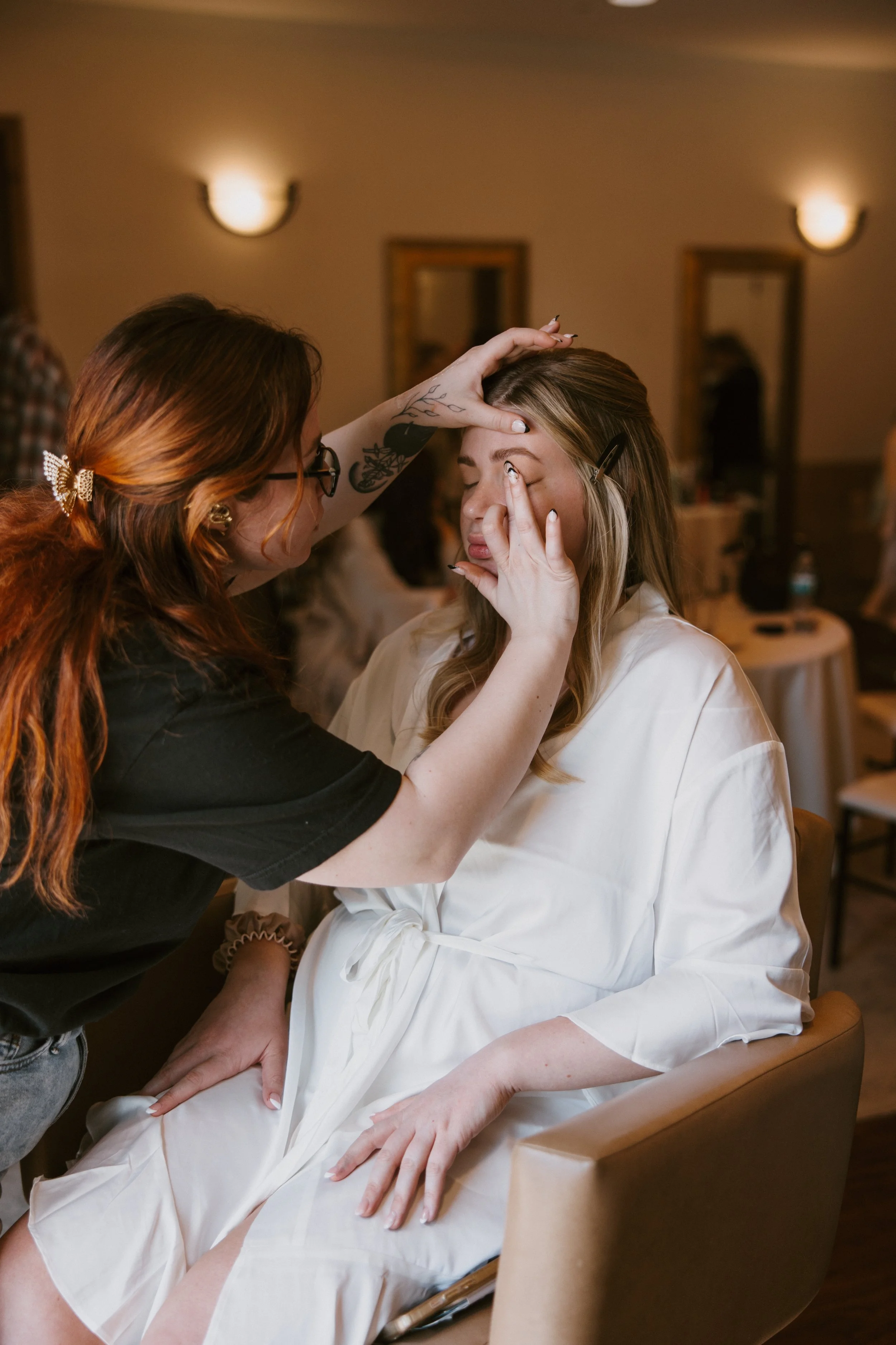 A woman receiving makeup application at a salon or event, sitting in a beige chair with a white robe, while a makeup artist stands beside her, touching up her eye makeup, in a warmly lit room with mirrors and people in the background.