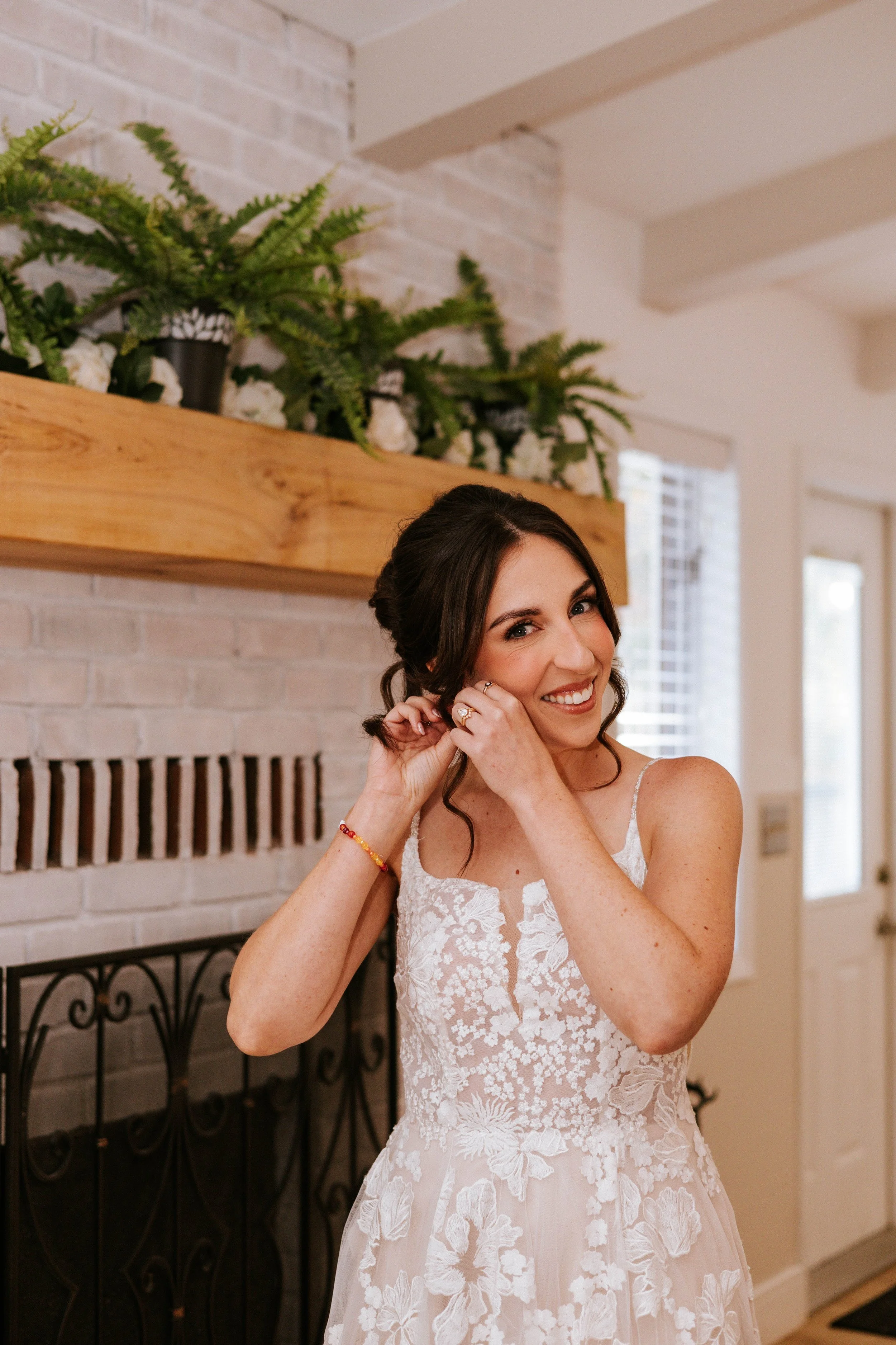 A woman in a wedding dress smiling and holding her hair in a room with a brick fireplace and a wooden mantel with potted plants.