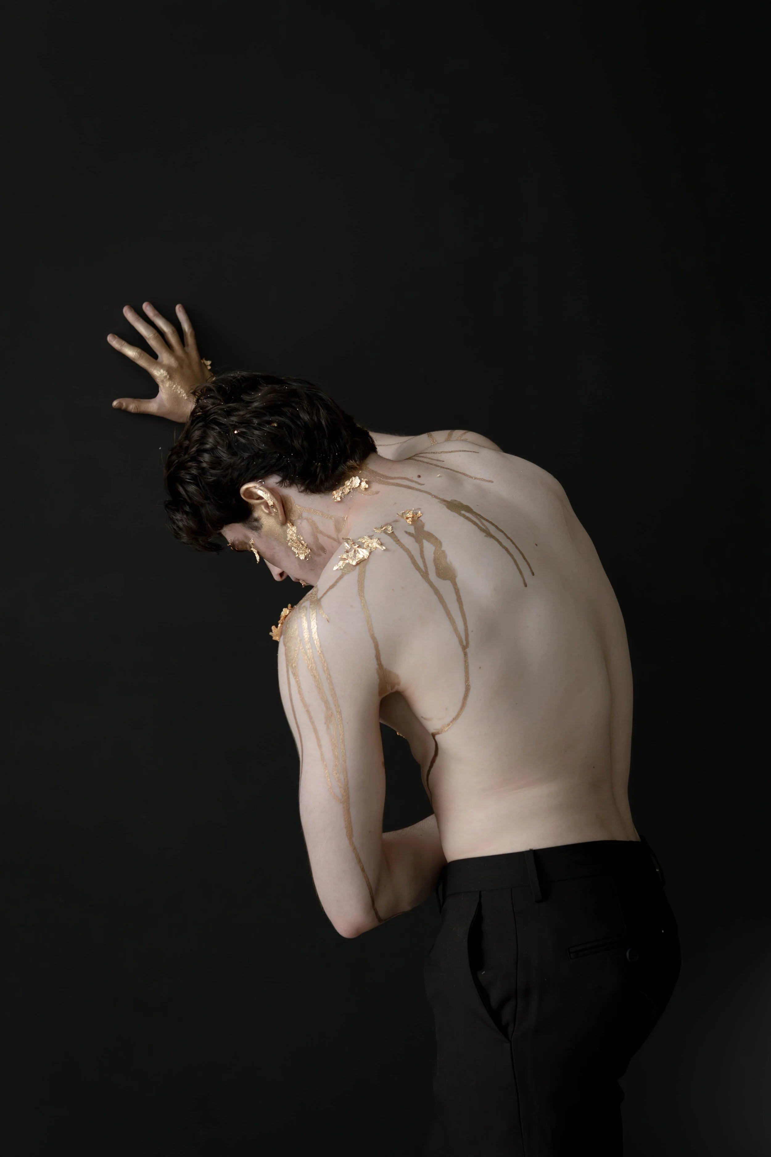 A person with short dark hair is topless against a black background, with gold paint splashed on their face, neck, arm, and back, and the person is wearing black pants.