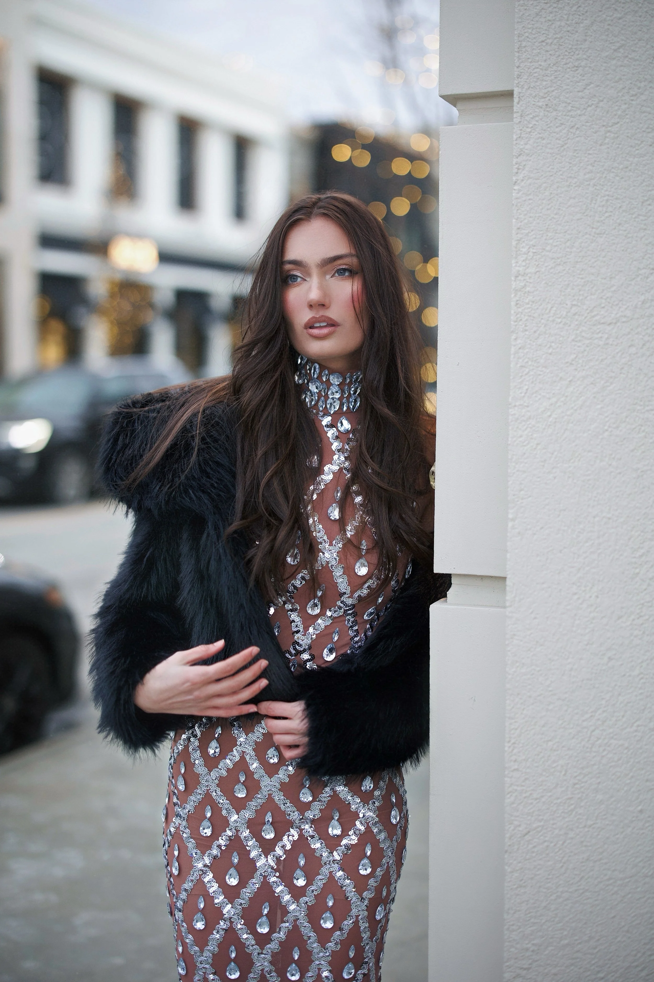 A woman with long dark hair, wearing a sheer dress with a diamond pattern, a large crystal necklace, and a black fur coat, peeks out from behind a white wall on a city street with blurred buildings and lights in the background.