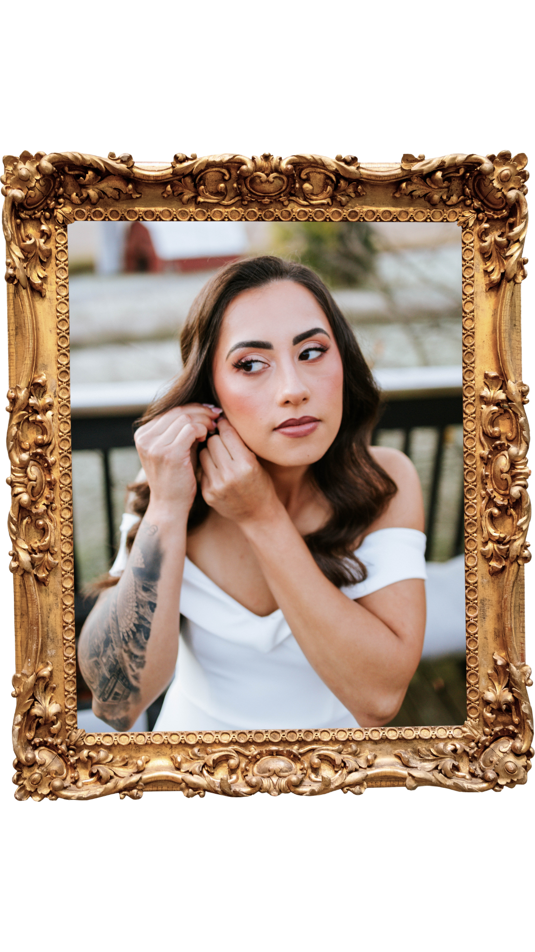 A woman with dark hair and makeup wearing a white off-shoulder dress, putting on earrings, sitting outdoors with a background of a pond and trees, framed by ornate gold decorative frame.