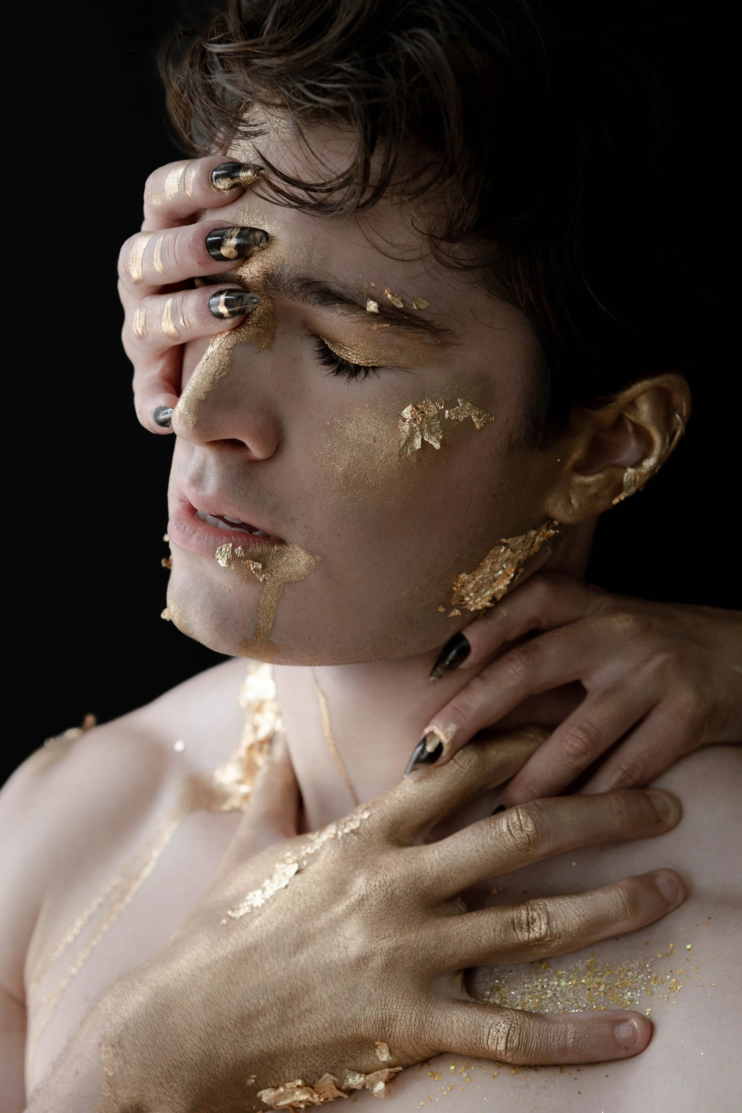 A person with short, curly hair is posed with one hand covering part of their face. Their eyes are closed, and their face and hand are decorated with gold leaf and metallic gold makeup, creating an artistic, surreal look.